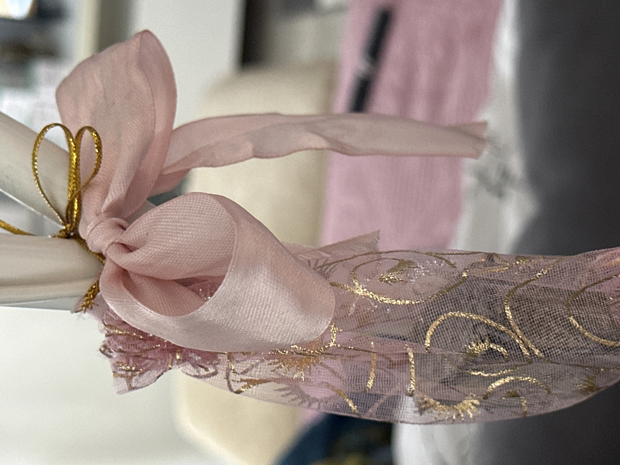 The image shows a close-up of a pink ribbon tied in a bow around a white cylindrical object. The ribbon is made of satin and has a gold, shimmering thread woven into it. A sheer pink fabric with gold filigree patterns is draped around the base of the object and tied with the ribbon. The background is blurred and shows a pink surface, possibly a bed or a tablecloth, and a dark gray surface, suggesting an indoor setting. There are no people or overt actions depicted. The overall impression is one of decoration or embellishment. The specific location of Nevinnomyssk, Russia is not visually represented in the image.