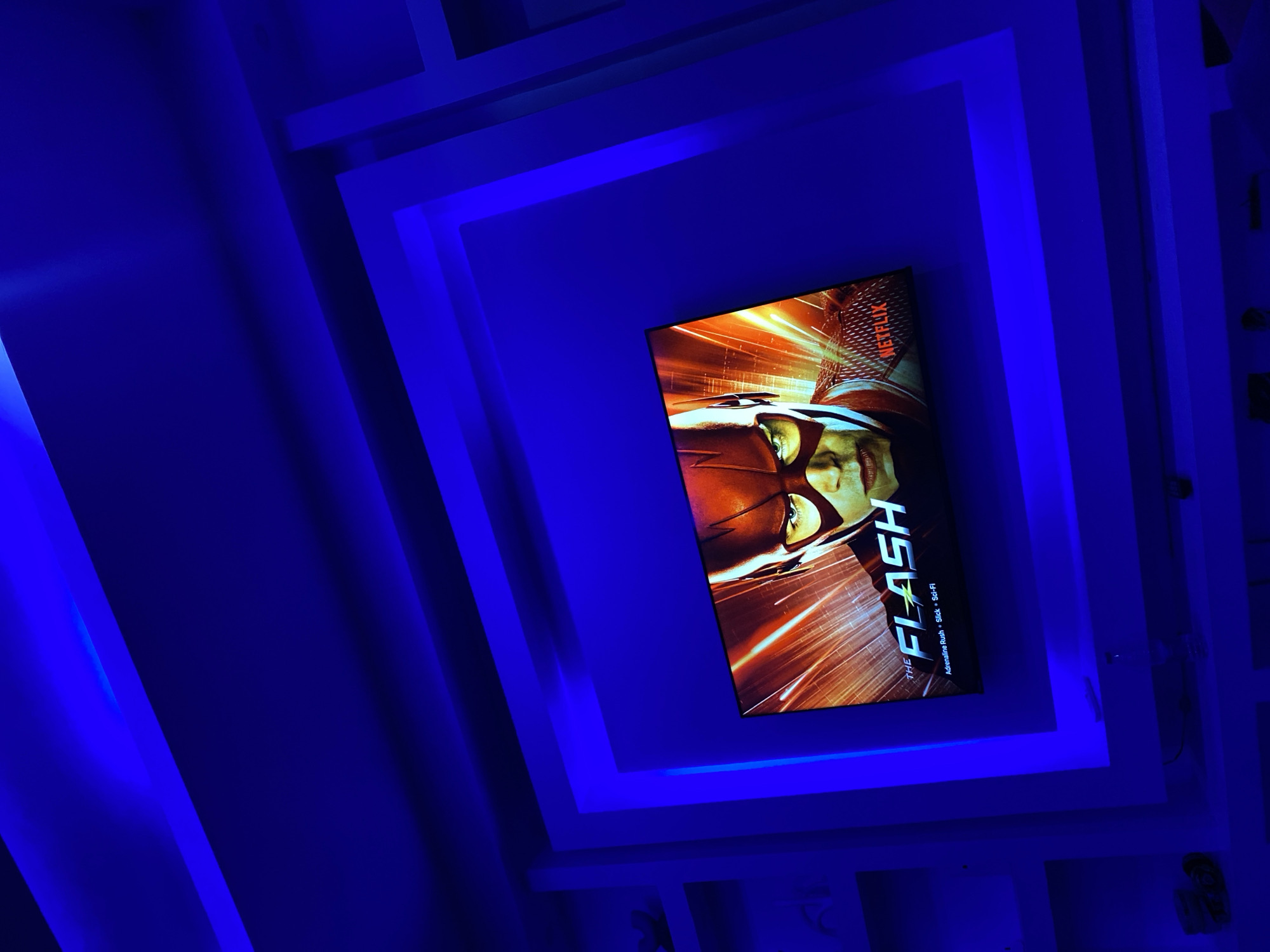 A low-angle shot depicts a television screen mounted in a recess of a ceiling featuring an intricate, multi-layered square design with integrated blue LED lighting. The television displays promotional art for the television show "The Flash" available on Netflix, described as an action/adventure/sci-fi series. The scene is dimly lit, emphasizing the blue glow emanating from the ceiling's recessed lighting. The location is not discernible from the image.