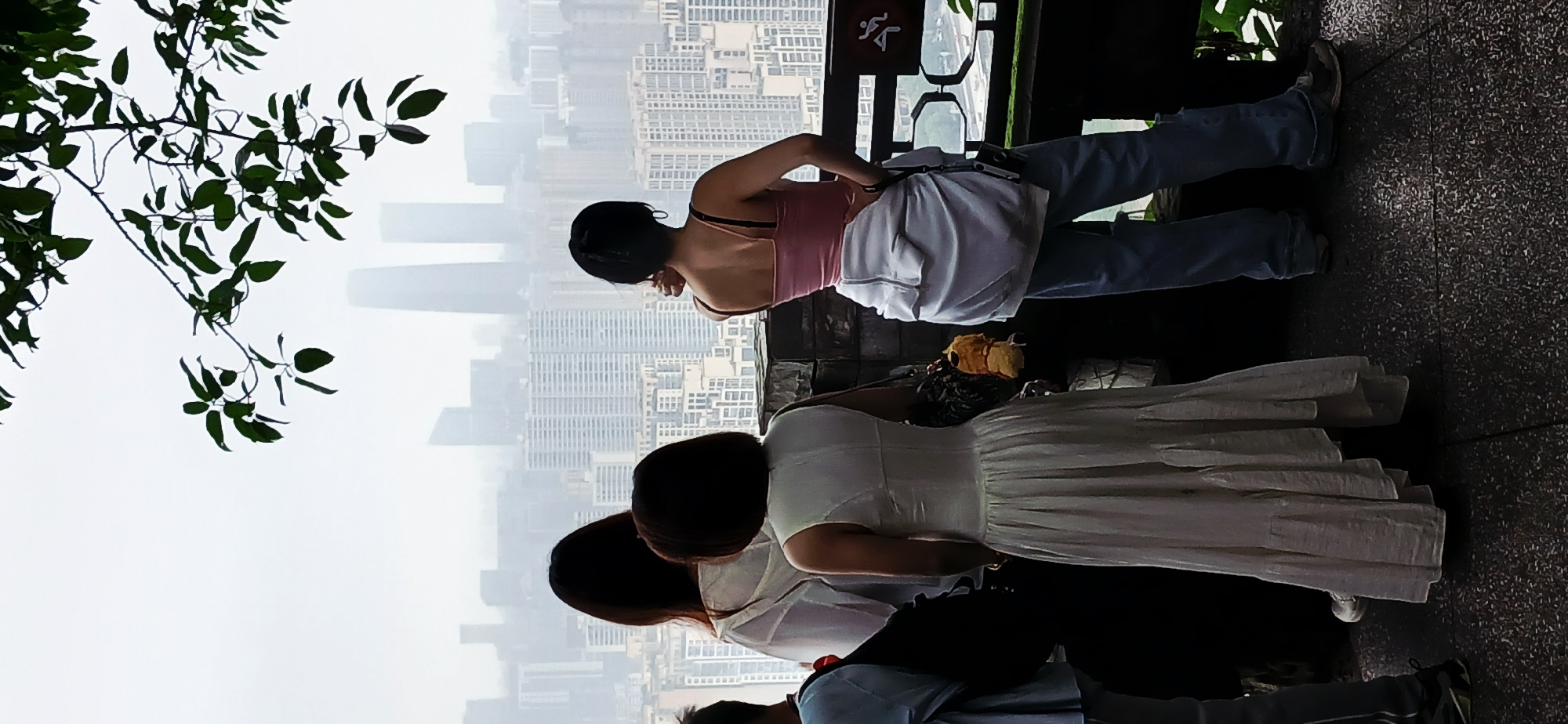 The photograph depicts a group of people standing on an elevated platform, possibly a viewing point, overlooking a city skyline shrouded in mist. The location appears to be in Yuzhong District, China, judging by the architecture of the buildings.
The people are positioned at varying distances from the camera. The individual closest to the camera is wearing a white dress. Others are clad in casual attire. They appear to be looking out at the city skyline.
The background features numerous high-rise buildings, barely visible through the mist. There are some trees and foliage visible in the top left corner of the image. A railing or barrier is visible, separating the people from the cityscape. The image suggests an overcast day with limited visibility.