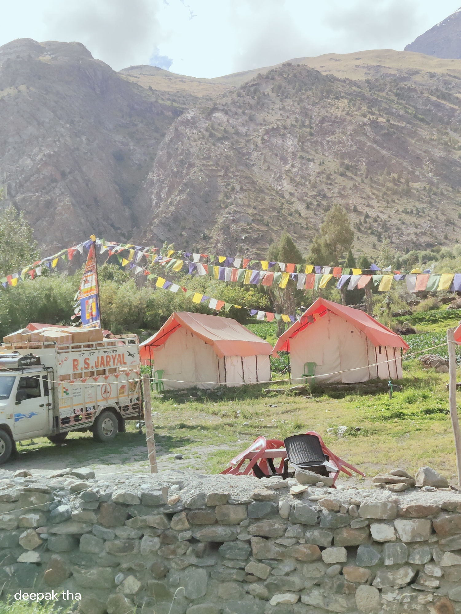 The image depicts a scenic view in Gemur, India, featuring a campsite with tents set against a backdrop of rugged mountains. In the foreground, a stone wall runs horizontally, with a truck laden with goods parked to the left. The truck displays "R.S. JARYAL" on its side. Two canvas tents with orange roofs are pitched in the middle ground, with prayer flags strung between poles above them. The terrain is characterized by dry, rocky slopes and sparse vegetation, typical of a mountainous region. Several red plastic chairs and a black woven chair are stacked near the tents. The sky is overcast with scattered clouds. The bottom left corner of the image contains the text "deepak tha".
