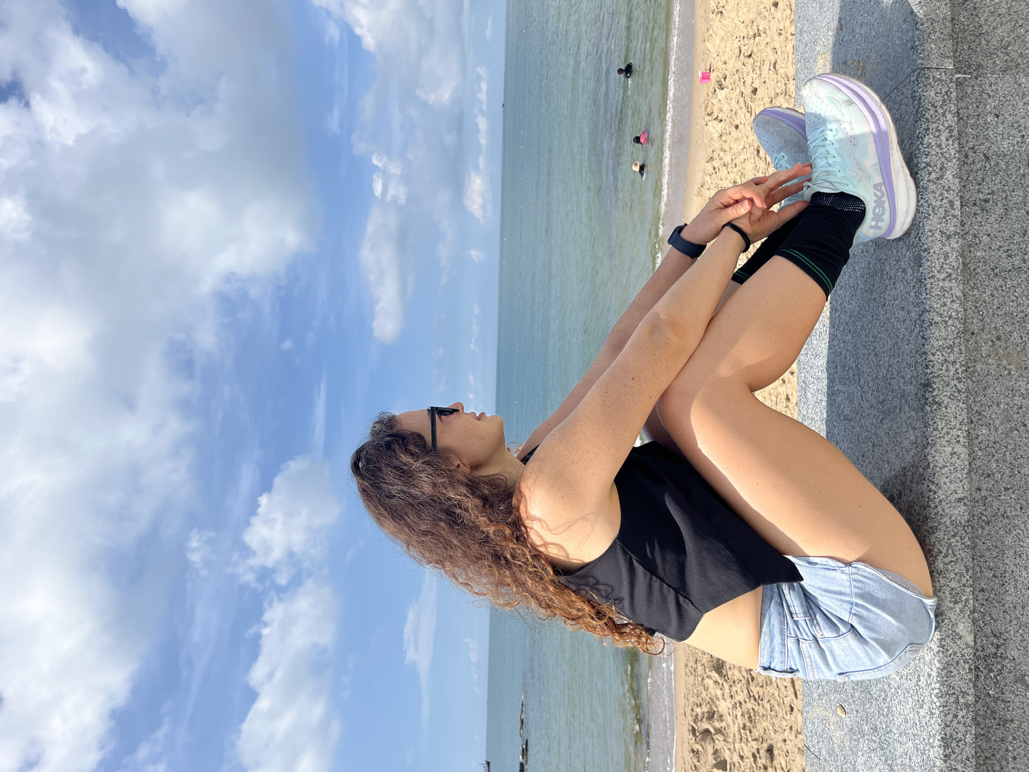 A young woman with curly brown hair and sunglasses is sitting on a grey stone wall, facing the ocean. She is wearing a black tank top, denim shorts, black socks, and light blue and purple athletic shoes. Her arms are resting on her knees. The background shows a sandy beach, the ocean, and a blue sky with white clouds. In the water, there are a few people swimming. The scene is located in Phan Thiết, Vietnam, likely along the coast.