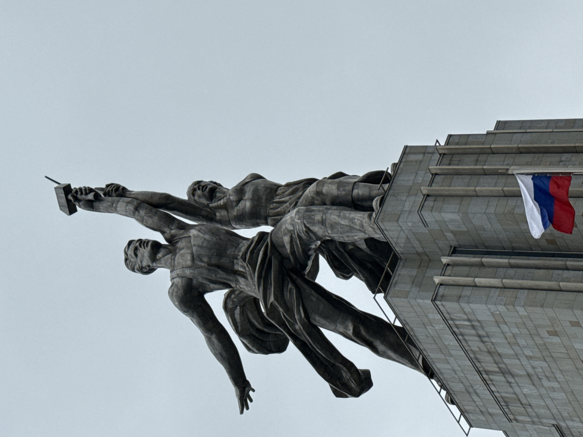The image shows the "Worker and Kolkhoz Woman" monument in Moscow, Russia. The statue depicts two figures made of metal: a male worker holding a hammer aloft and a female kolkhoz worker holding a sickle. They are posed as if striding forward together, symbolizing the unity and progress of the Soviet Union. The monument is mounted atop a building with a Russian flag hanging from it. The background is a cloudy sky.