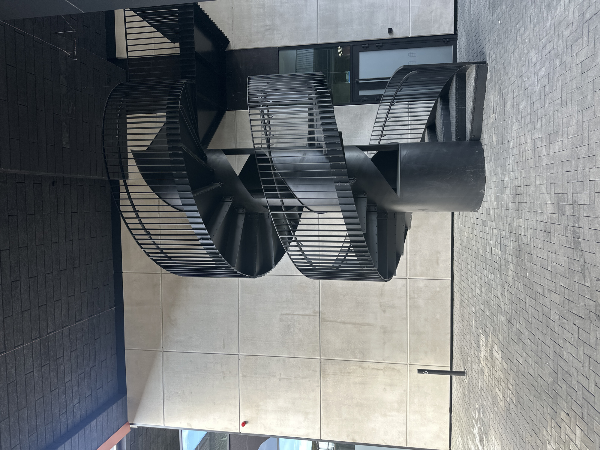 The image depicts a modern, outdoor spiral staircase with a black metal railing and steps, set against a textured concrete wall. The staircase is centrally located in the frame and curves upwards and to the right. The ground is paved with grey interlocking bricks arranged in a herringbone pattern. Above the concrete wall, a dark, textured ceiling is visible, with some suspended wires and lighting fixtures. To the right of the staircase, a glass-paneled door with a black frame is set into the concrete wall, leading into a building. The overall scene suggests an urban, contemporary architectural setting, likely a public space or the exterior of a modern building. The presence of a single black pole with a number "6" on it near the bottom left of the image suggests a possible identification or marking within the area. The lighting indicates it is daytime, with shadows cast on the paved ground, suggesting the sun is to the left and slightly behind the viewer. There are no people visible in the image. The architecture and materials suggest a minimalist, industrial aesthetic.