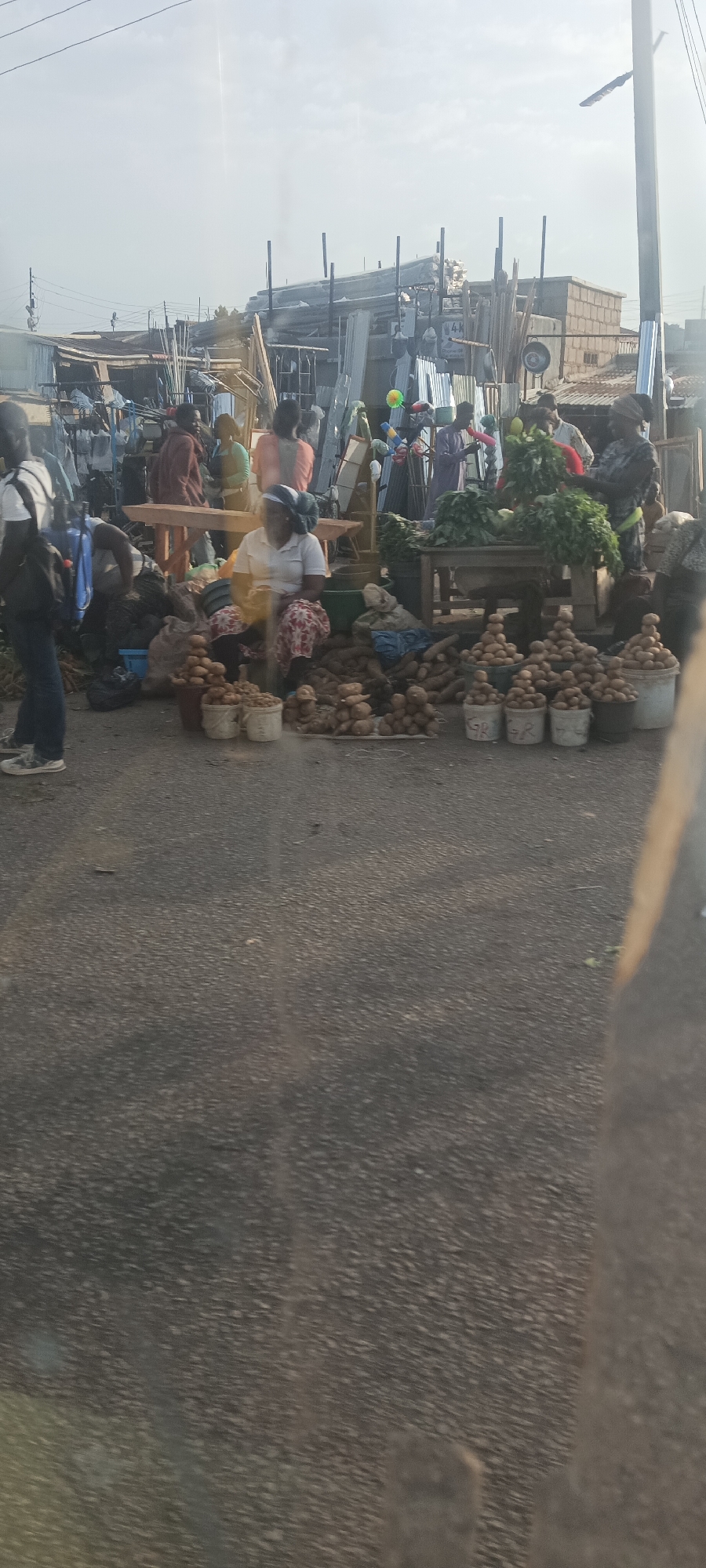 This media file captures an active outdoor market scene, likely in Gura Top, Nigeria, as suggested by the context. The setting is a paved street or roadside, with numerous vendors and shoppers engaged in daily commerce.
In the foreground, a prominent female vendor is seated on the ground, wearing a white top, a patterned skirt, and a headwrap. She is surrounded by a large array of fresh produce. Abundant piles of root vegetables, possibly yams or similar tubers, are laid directly on the asphalt. Neatly arranged in several white buckets are stacks of smaller, round vegetables, which appear to be potatoes. Some buckets display the text "GR". Adjacent to her, a simple wooden table holds generous bunches of leafy green vegetables for sale.
Several other individuals are visible throughout the scene. To the left, a person carrying a backpack walks away from the viewer. Other people are seen standing, conversing, or tending to their stalls in the background. The structures lining the market area are informal, constructed with corrugated metal sheets and basic building materials, hinting at the sale of hardware or construction supplies, with visible stacks of metal and beams. A sign in the background clearly shows "4K". Colorful items, possibly toys or household goods, are also displayed on racks in the background.
The lighting indicates a bright day, likely late morning or early afternoon, under a clear or partly cloudy sky. The overall atmosphere is one of bustling market activity, showcasing the trade of fresh agricultural produce and other goods in a vibrant local setting.