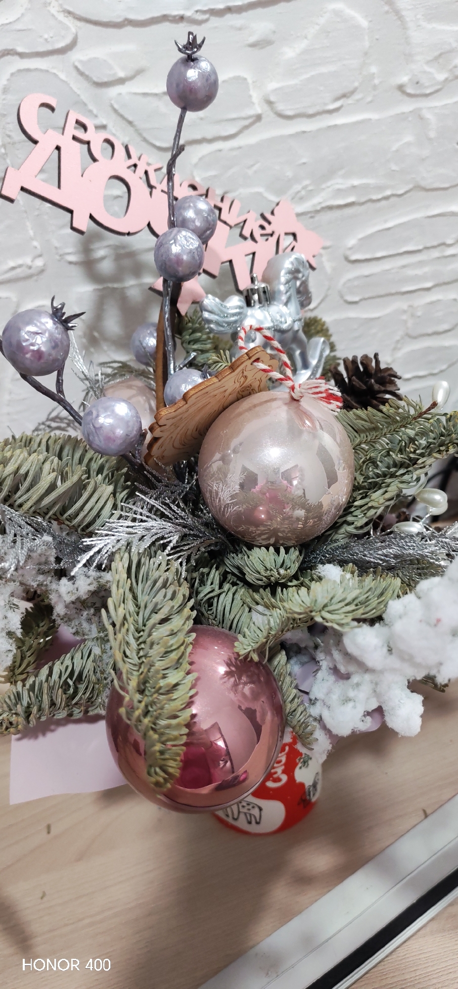 The image depicts a Christmas or New Year's centerpiece arrangement. It contains pine branches (some with faux snow), silver-colored decorative twigs with spherical ornaments, two pink/rose-colored Christmas ornaments, a wooden tag, a silver-colored horse ornament, and a small pinecone. Pink wooden letters are in the background and are likely part of a New Year's greeting in Russian. The centerpiece is set against a white brick-like background. The inscription "HONOR 400" in the lower left corner suggests the image was taken by an Honor 400 device. There is nothing in the image that indicates it was taken in the city of Noviy, Russia.