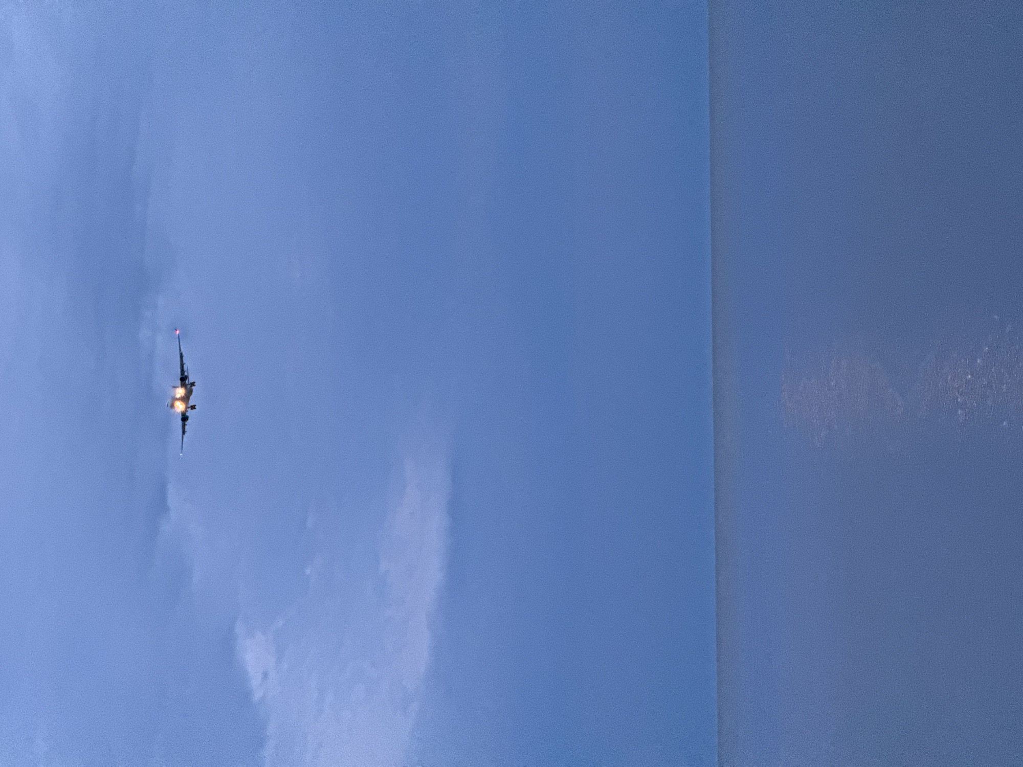 The image displays an airplane in flight against a deep blue, twilight sky, with the given location context of Sochi, Russia. The aircraft is positioned in the upper left quadrant, viewed from below. Its engines are brightly illuminated with a warm orange glow, indicating either dusk or early evening. A small red light is visible near the tail of the plane. The sky is predominantly clear, showing subtle shifts in blue hues and a few faint, wispy clouds. A prominent vertical boundary divides the image on the right, revealing a solid, muted blue-gray surface. This suggests the photograph was taken from an indoor vantage point, possibly through or past a window frame or against an interior wall. No people or visible text are present in the image. The scene captures the solitary presence of the aircraft against a calm, evening sky.