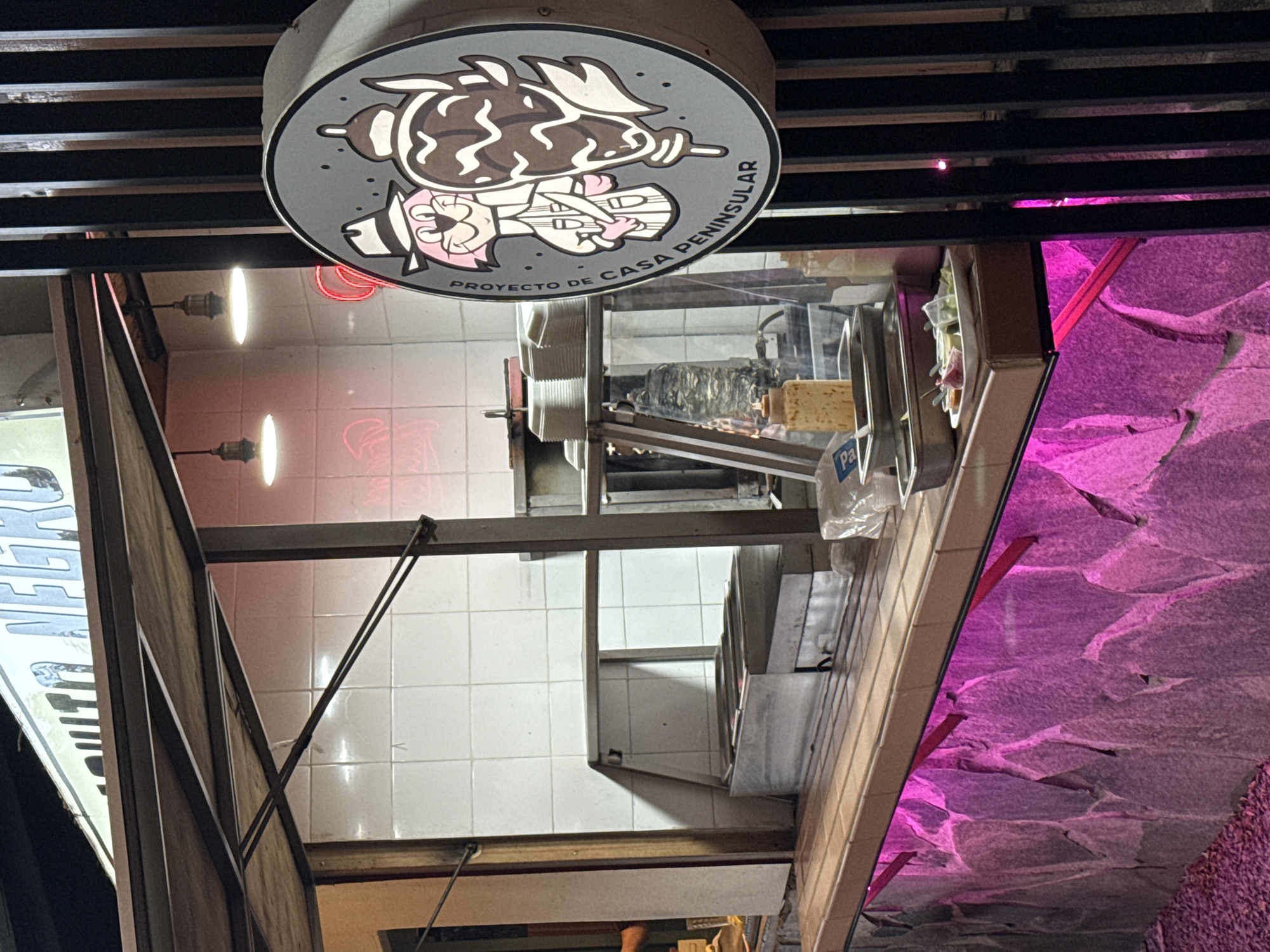 The image displays an illuminated outdoor food stall at night, located in Puebla, Mexico. The stall features a wooden-tiled service counter set atop a rough-cut stone base, which is underlit by pink LED strips. The interior rear wall of the stall is clad with white square tiles. Visible behind the counter are various stainless steel commercial kitchen appliances, including open food warmers and a vertical rotisserie (trompo) with a large conical stack of meat. Two industrial-style pendant lights hang from the overhead canopy, providing primary illumination to the workspace.
On the service counter, multiple stainless steel trays are present, along with squeeze bottles containing condiments (one yellowish, one white). A white rectangular tray holds small plastic cups filled with diced onions, cilantro, lime wedges, and both red and green sauces. To the right of the counter, a circular, backlit sign is affixed to a dark, vertically slatted structure. This sign depicts a cartoon pink, bipedal, cat-like character wearing a fedora and trench coat, standing next to a stylized vertical rotisserie. The text "PROYECTO DE CASA PENINSULAR" encircles the central graphic. An overhead sign, partially visible at the top of the frame, reads "BURRITO NEGRO." No individuals are present in the scene. The ground immediately in front of the stall is covered with small gravel.