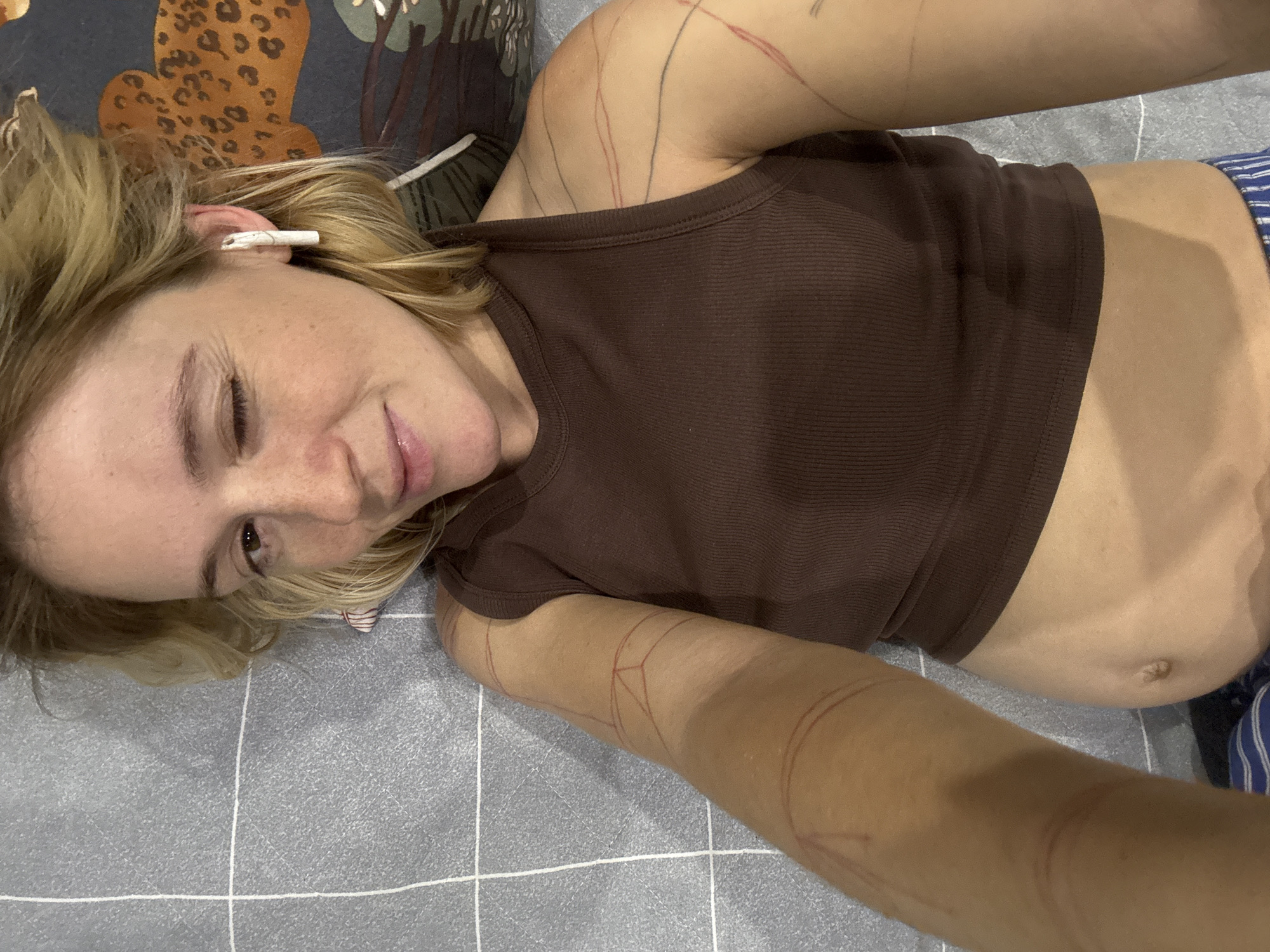 A woman lies on a grey, checkered surface, possibly a bed or mat. She is wearing a brown ribbed tank top and has blonde hair. She is looking towards the camera with one eye open and the other closed in a wink. There are red markings on her arms that appear to be drawn or tattooed designs. She is also wearing white earbuds. The background shows a pattern with what appears to be leopard print. The image does not provide any information about the location in the city of Nizhny Novgorod, Russia.
