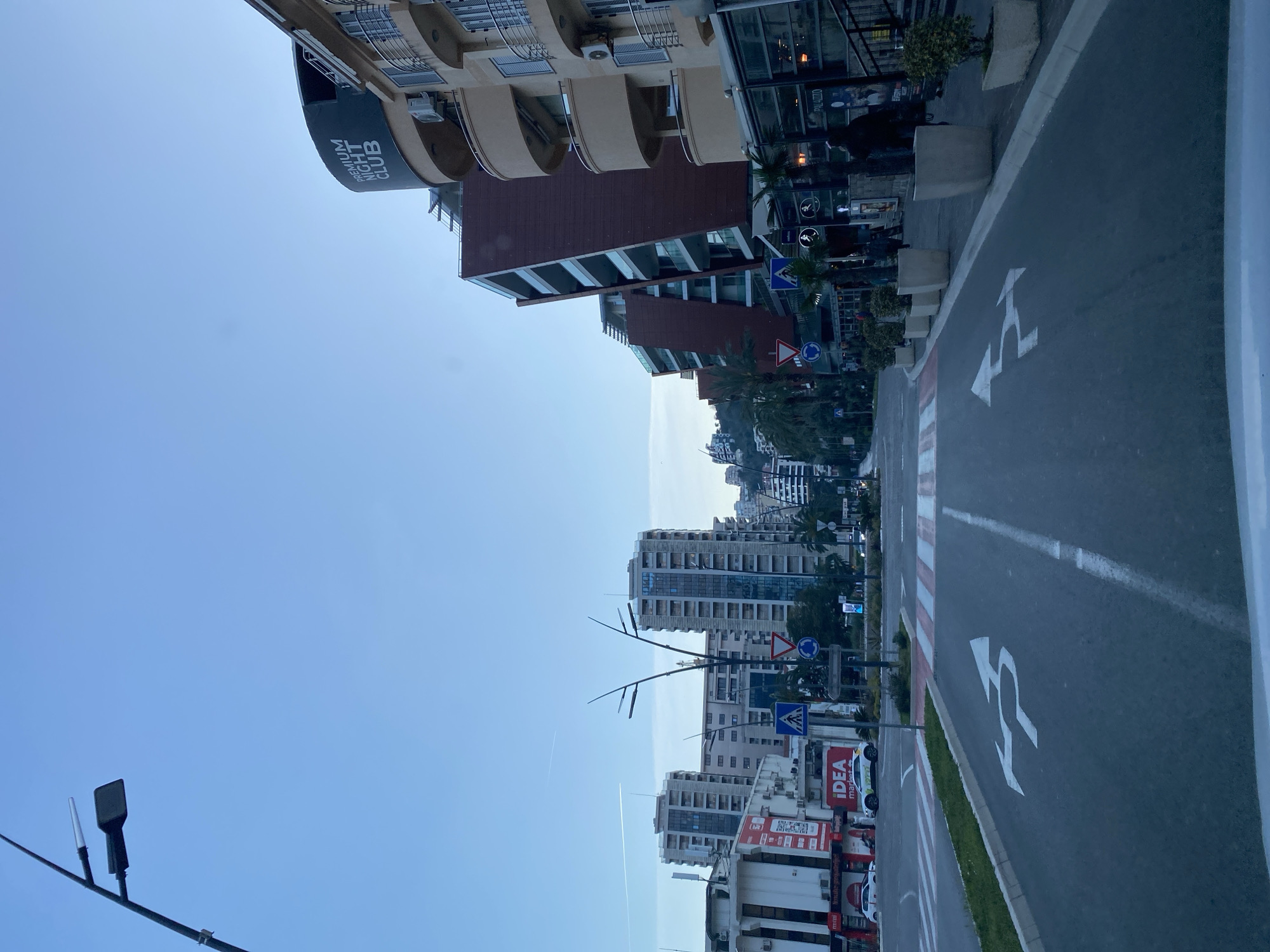 The image depicts a street scene in Budva, Montenegro. The camera is angled upward, capturing buildings, street signs, and the sky. Several buildings are visible along the street, including a structure with a "PREMIUM NIGHT CLUB" sign. There's a multi-story building with balconies and a modern architectural style. The street has painted lane markings and directional arrows. The street also has an IDEA market. The sky is mostly clear with a slight trace of an aircraft contrail. A lamp post is visible in the lower left corner. Trees and vegetation are present along the street. The buildings vary in height and design, showcasing a mix of residential and commercial structures. The city extends into the background.