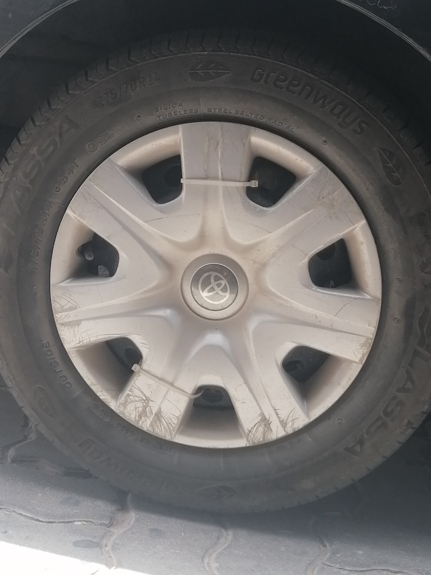 An extreme close-up shot focuses on the wheel of a car parked on a cobblestone surface. The tire, a Greenway brand, is marked with "175/70R14" and "OUTSIDE." A silver hubcap with the Toyota logo is visible in the center. White zip ties are used to secure a portion of the tire to the hubcap. The scene is likely a street or parking area in Essaouira, Morocco, given the cobblestone texture of the ground.