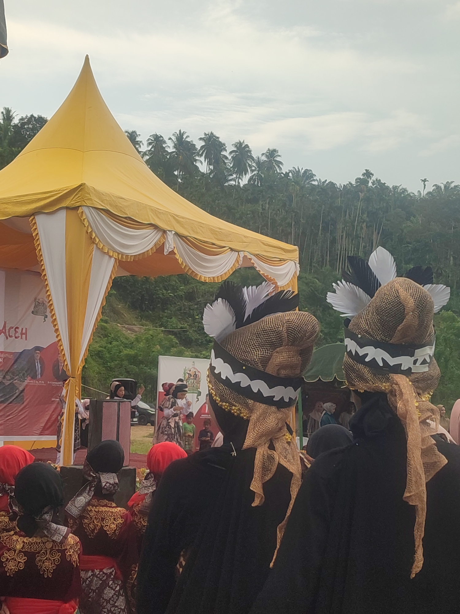 This image depicts an outdoor cultural event under a yellow and white tent in the city of Langkuta, Indonesia. In the foreground, two individuals are seen from behind, wearing elaborate headwear with black and white feathers, and woven hats decorated with gold netting and ribbon. They are also adorned with gold beaded necklaces and tan netting draped over their heads and shoulders. Behind them, a crowd of people are gathered, many wearing traditional Indonesian attire, including richly embroidered red and black garments and headscarves in various colors like red and black. A stage area is visible under the tent, with a red banner displaying text and images, including a portrait of a man in a suit. In the middle ground, several children are performing or observing the event, one child in traditional attire is standing in front of a raised platform. A dark vehicle is parked behind the stage area. The background shows a lush, densely forested hillside with numerous palm trees, suggesting a rural or semi-rural setting. The sky is overcast with a few scattered clouds.