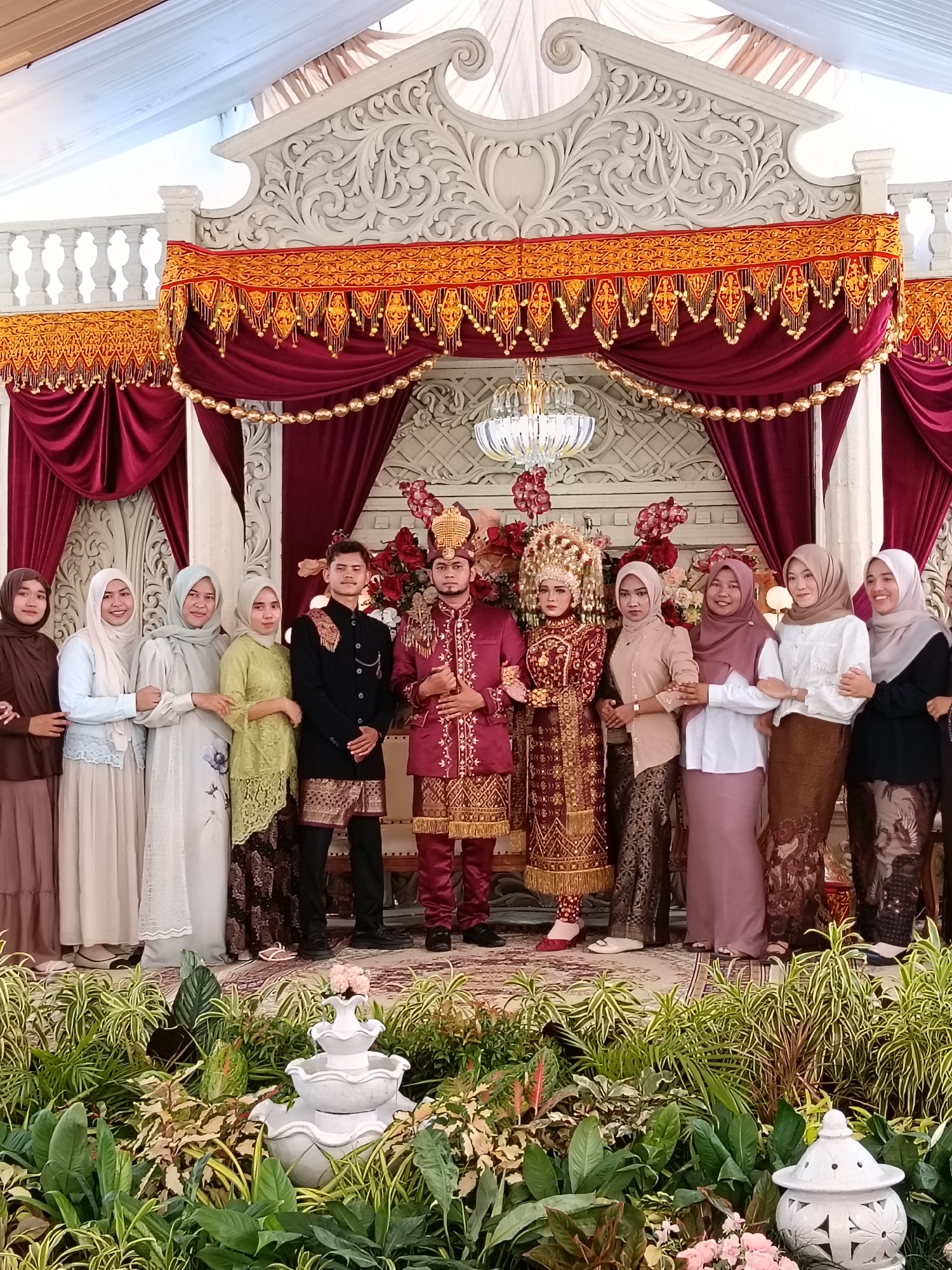 The image shows a group of people standing on a platform in front of a decorated backdrop, presumably at a wedding celebration. The platform is adorned with greenery and a small white fountain in the foreground.
In the center, a couple stands dressed in traditional attire. The man wears a red and gold outfit, while the woman wears a similarly colored and intricately designed dress with an elaborate headpiece. To their left and right, individuals, mostly women, are dressed in more contemporary attire, some wearing hijabs.
The backdrop consists of red velvet curtains, gold beaded garlands, and decorative carvings. A chandelier hangs above the couple. The setting appears to be indoors, possibly in a wedding hall.