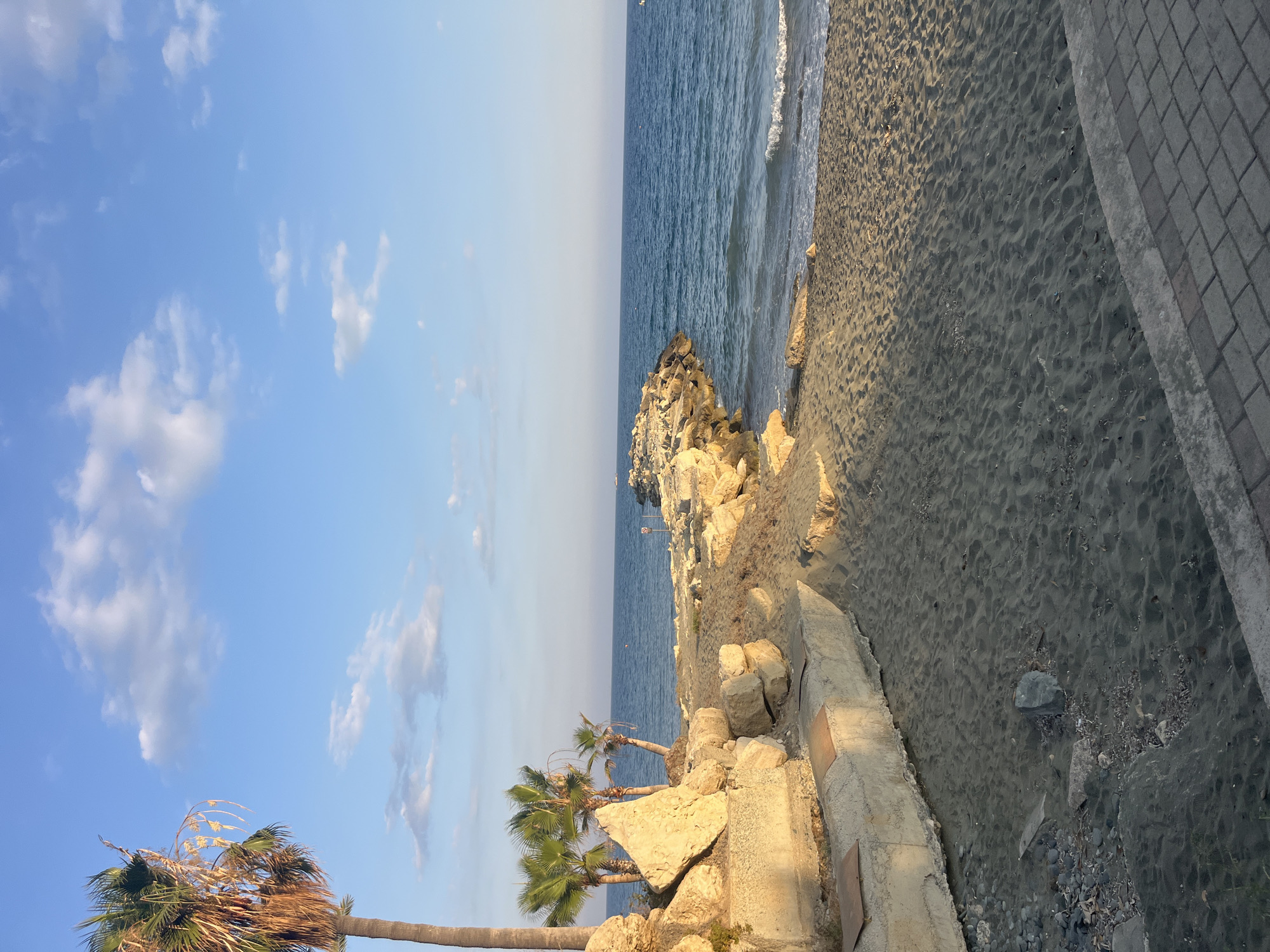 A long shot captures a sandy beach meeting the sea under a bright blue sky dotted with white clouds. A concrete structure runs along the bottom of the frame, leading to a sandy and rocky embankment on the left, where palm trees are visible. A rocky pier extends into the sea from the right side of the embankment. The sea is a deep blue, with gentle waves reaching the shore. The sun casts a warm glow on the sand and rocks. The overall scene suggests a tranquil coastal environment in Agios Tychona Community, Cyprus, during a clear day.