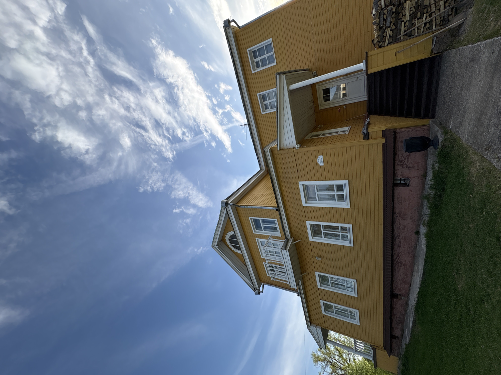 The image depicts a two-story, mustard-yellow wooden house against a backdrop of a blue sky streaked with white clouds. The house features white-framed windows of varying sizes on both floors, along with a small balcony and a covered entryway with steps leading up to the front door. A stack of firewood is visible next to the steps. Below the house, there is a foundation with a pinkish hue, and a patch of green grass in the foreground. The scene is likely located in a residential area. The house is identified by its architectural style as being located in Zavod Mikhailovskii, Russia.