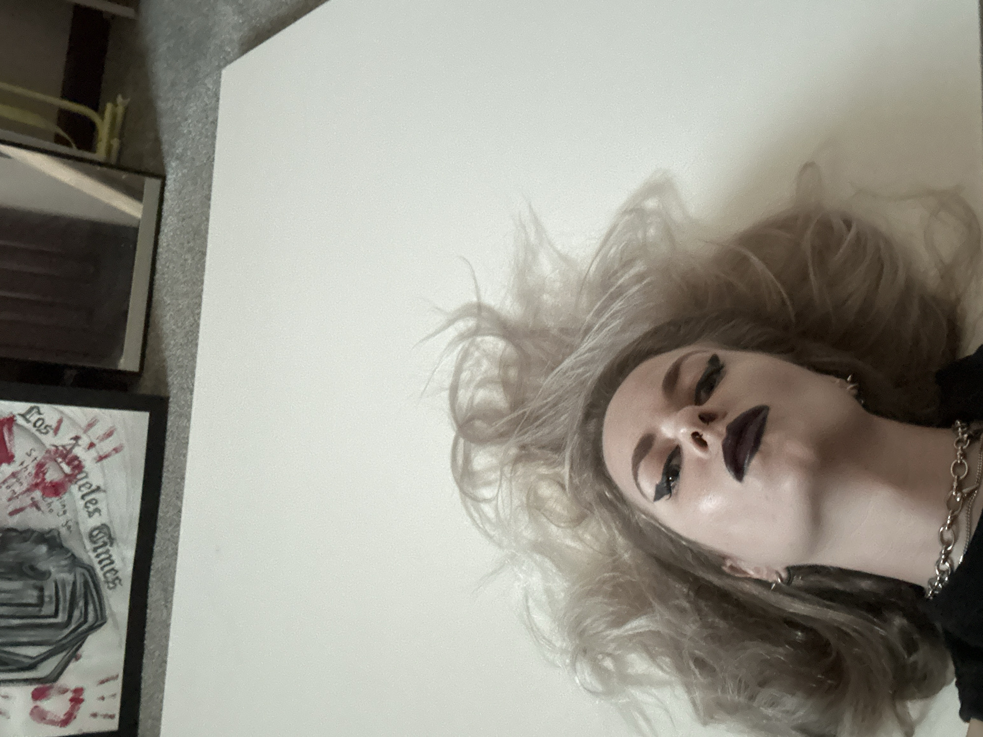 A photograph shows a light-skinned person with pale blonde hair and dark lipstick. The person is lying on a white surface with their head tilted to the right. The person is wearing a black shirt and a silver chain necklace. There is a framed poster with the words "Los Angeles Times" and drawings on the left side of the image, as well as a mirror partially visible above it. The photo's orientation suggests it was taken from an elevated position looking down. The location in Cape Coral, United States of America, is not determinable from the image.