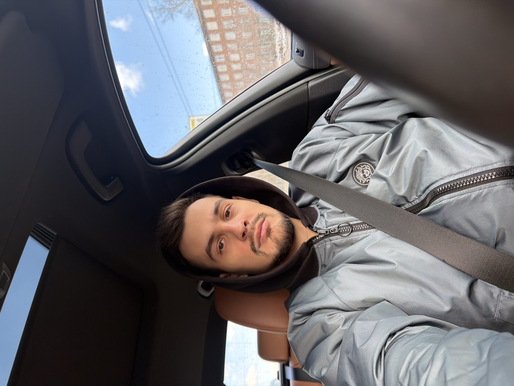 A selfie shows a man with dark hair and a beard wearing a gray jacket with a black hood and a seatbelt, presumably in a car. Above him is the open sunroof, revealing a view of a light blue sky with a few clouds, and a blurred building in the background. Based on the architectural style of the building visible through the sunroof, it's highly likely the location is in Krasnoyarsk, Russia. The interior of the car is mostly dark, with a glimpse of a tan leather headrest.