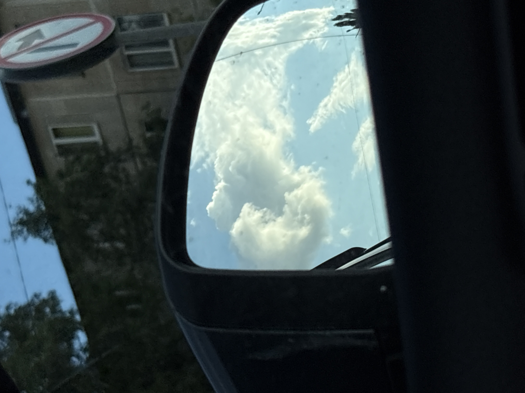This image is a photograph taken from inside a vehicle, looking at the side-view mirror. The mirror reflects a bright blue sky filled with large, puffy white clouds. To the left of the mirror, a dark car door frame is visible. In the background, outside the car and not reflected in the mirror, is a building with several windows, and a traffic sign with a red circle and a diagonal line crossing an arrow, indicating "No left turn." Trees are also visible in the background. The scene appears to be in an urban setting.