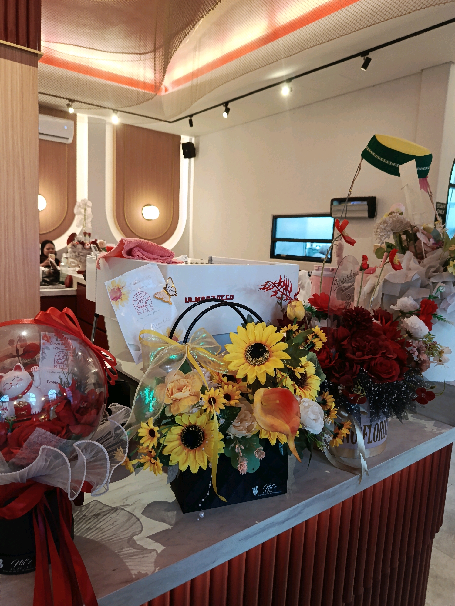 The image shows a counter in a retail establishment, possibly a cafe or flower shop, with several bouquets and decorative items arranged on its surface. In the background, there is an air conditioning unit mounted high on the wall, and two wall-mounted lights with a warm glow. The ceiling features a decorative netting structure with orange lighting.
In the foreground, a large clear balloon filled with red artificial flowers and a white maneki-neko (beckoning cat) figurine is prominently displayed. Next to it, a basket of yellow and red artificial flowers sits on the counter, adorned with a shimmery ribbon. To the right of this arrangement, another bouquet features red roses and various other flowers. Part of a white box with the "La Marzocco" logo is visible behind the flower arrangements. A card with sunflowers and a butterfly sticker is also placed on the counter. The counter itself is made of a light-colored material with a textured, reddish-orange base.
In the middle ground, a woman is visible through a doorway in the background, seated at a table and looking towards the left. Another person is partially visible behind her. A window with a bluish tint is seen above the counter. A yellow and green cap is perched on a decorative item in the background. A sign with "STAFF ONLY" text is also visible in the far right background.
The overall impression is one of a brightly lit, modern commercial space decorated with floral arrangements, likely for a special occasion. The text on the white box and the card suggest that this might be a celebration or a gift-giving event. The presence of the "La Marzocco" logo indicates a connection to coffee, possibly a cafe. However, without further context, the specific location in Kudus, Indonesia, cannot be determined solely from the visual information provided in the image.
