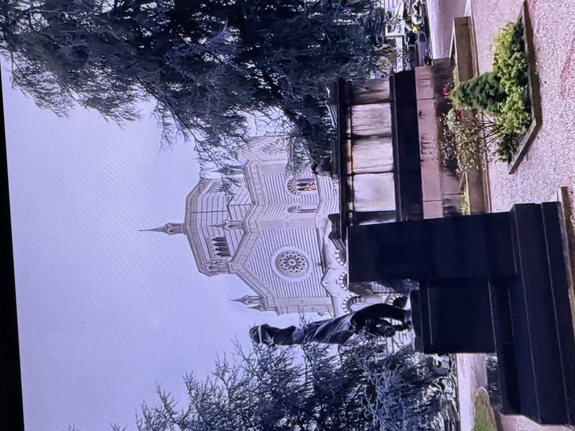 This image captures a cemetery scene with a large, ornate building resembling a church or mausoleum in the background. In the foreground, a dark stone monument with a sculpted figure is visible. There are also several trees and manicured garden beds within the cemetery. The sky is overcast and the overall lighting suggests a somber or cloudy day. There is no indication that this location is in Dubai, United Arab Emirates.