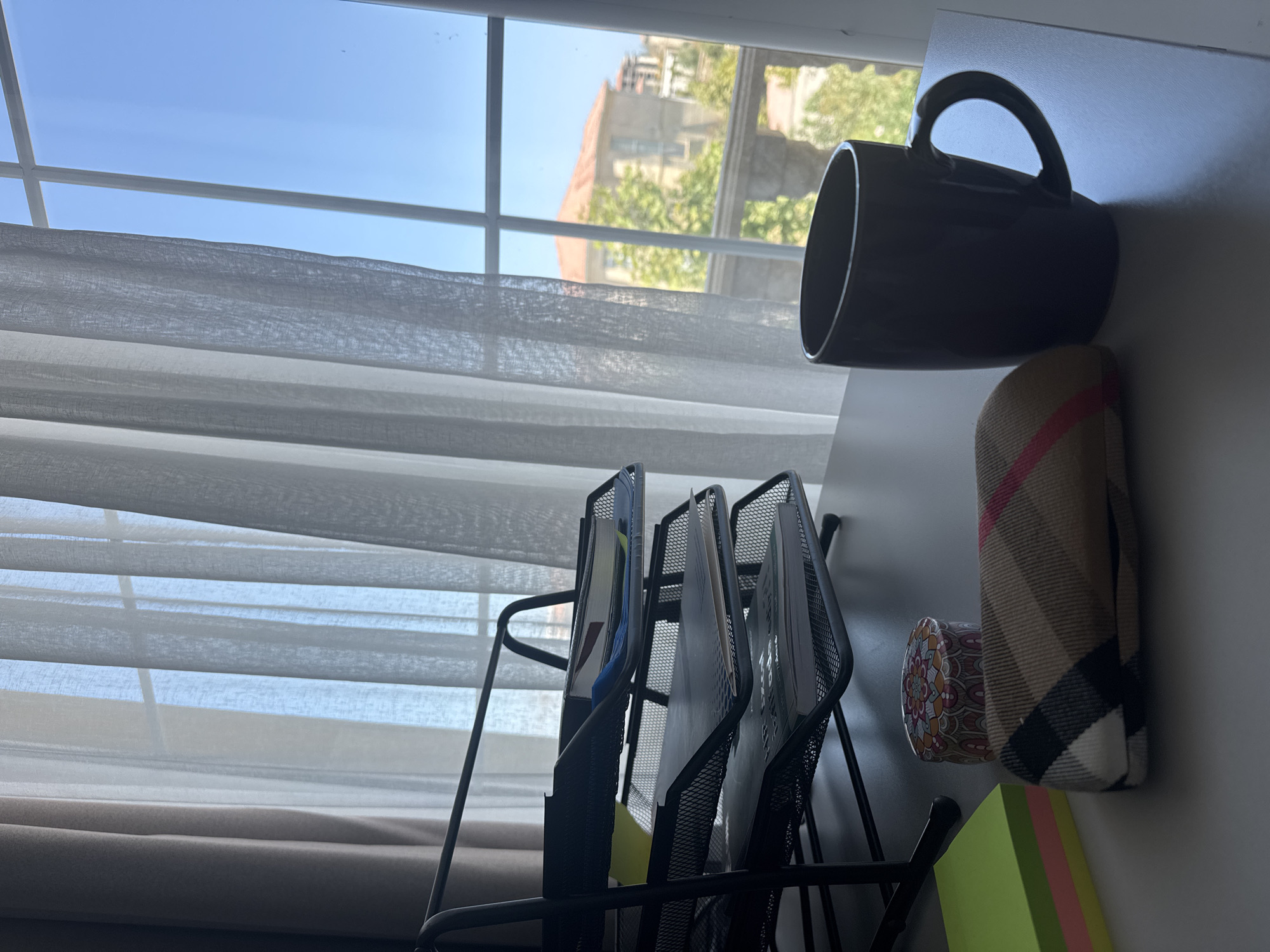 A desk is set up by a window with a view of buildings and trees in the Sabunchu Raion of Azerbaijan. The desk holds a black metal document organizer filled with papers, a stack of colorful sticky notes, a small round patterned tin, a fabric-covered glasses case, and a dark mug. The window is covered with sheer white curtains, allowing natural light to illuminate the scene. Through the window, a clear blue sky is visible, along with the rooftops and facades of residential buildings, and lush green trees.