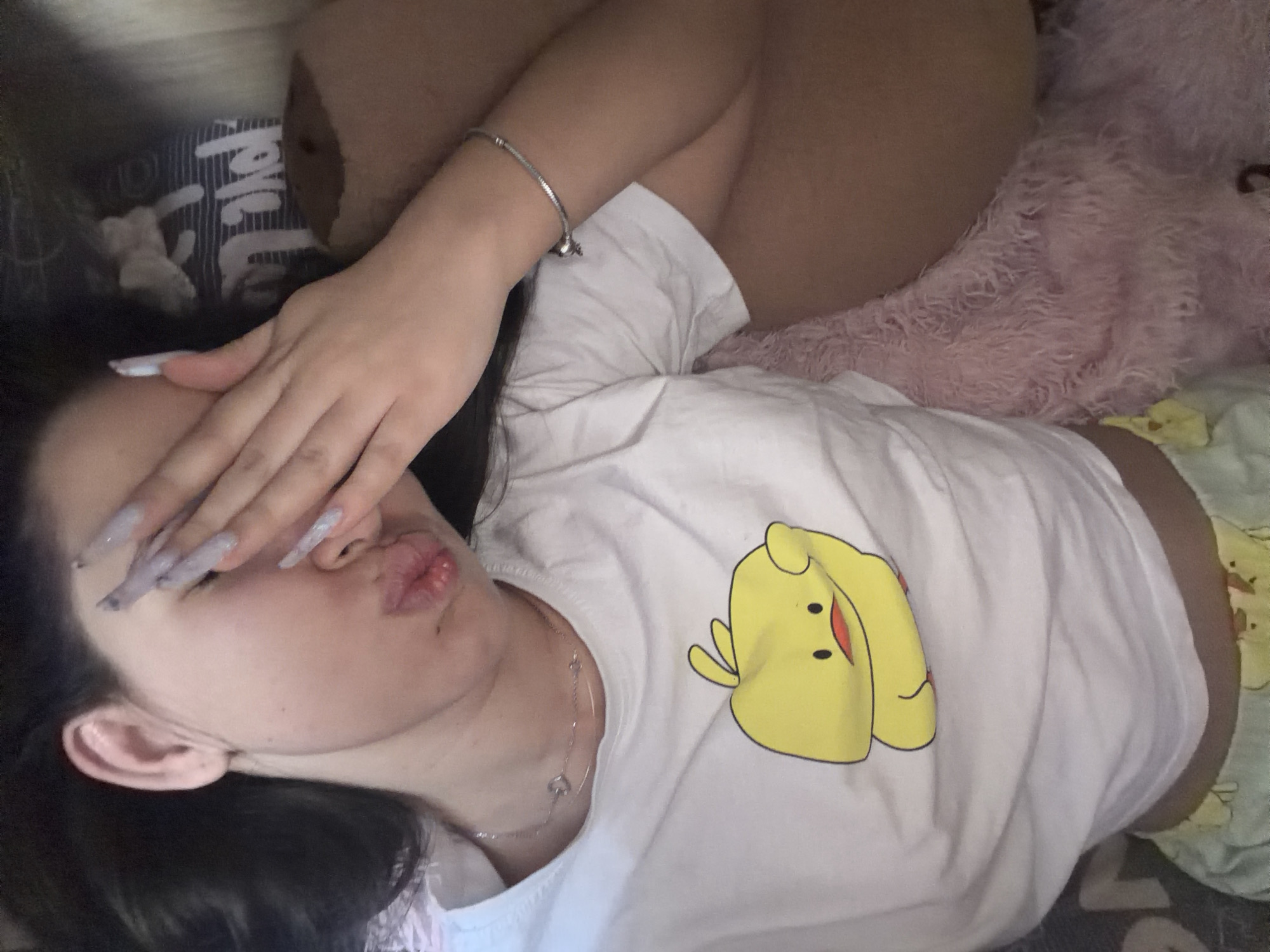 A person is lying on a bed with a pink stuffed animal to their right. They are wearing a white t-shirt with a yellow cartoon image. Their left hand is covering their face. A silver bracelet is on the person's right wrist. The location is not discernible from the image.