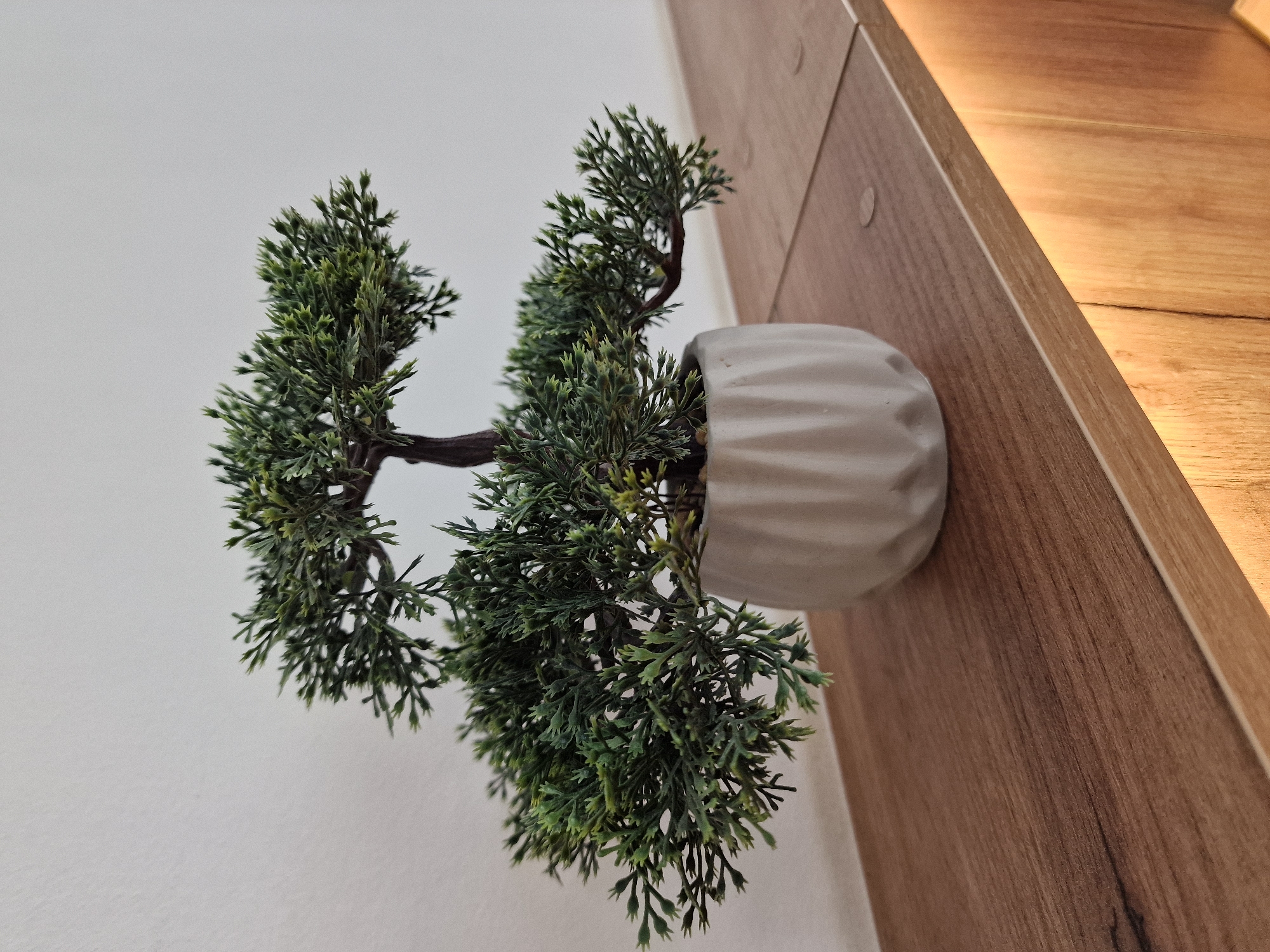 A small, artificial bonsai tree with dark green foliage sits in a light gray textured pot on a wooden shelf. The pot has a fluted design around its base. The shelf is made of wood with a visible grain and is lit from below by warm yellow lights, casting an orange glow on the vertical wooden panels beneath it. The background is a plain, light-colored wall. The scene is indoors, suggesting it is likely a decorative object in a home or office setting.