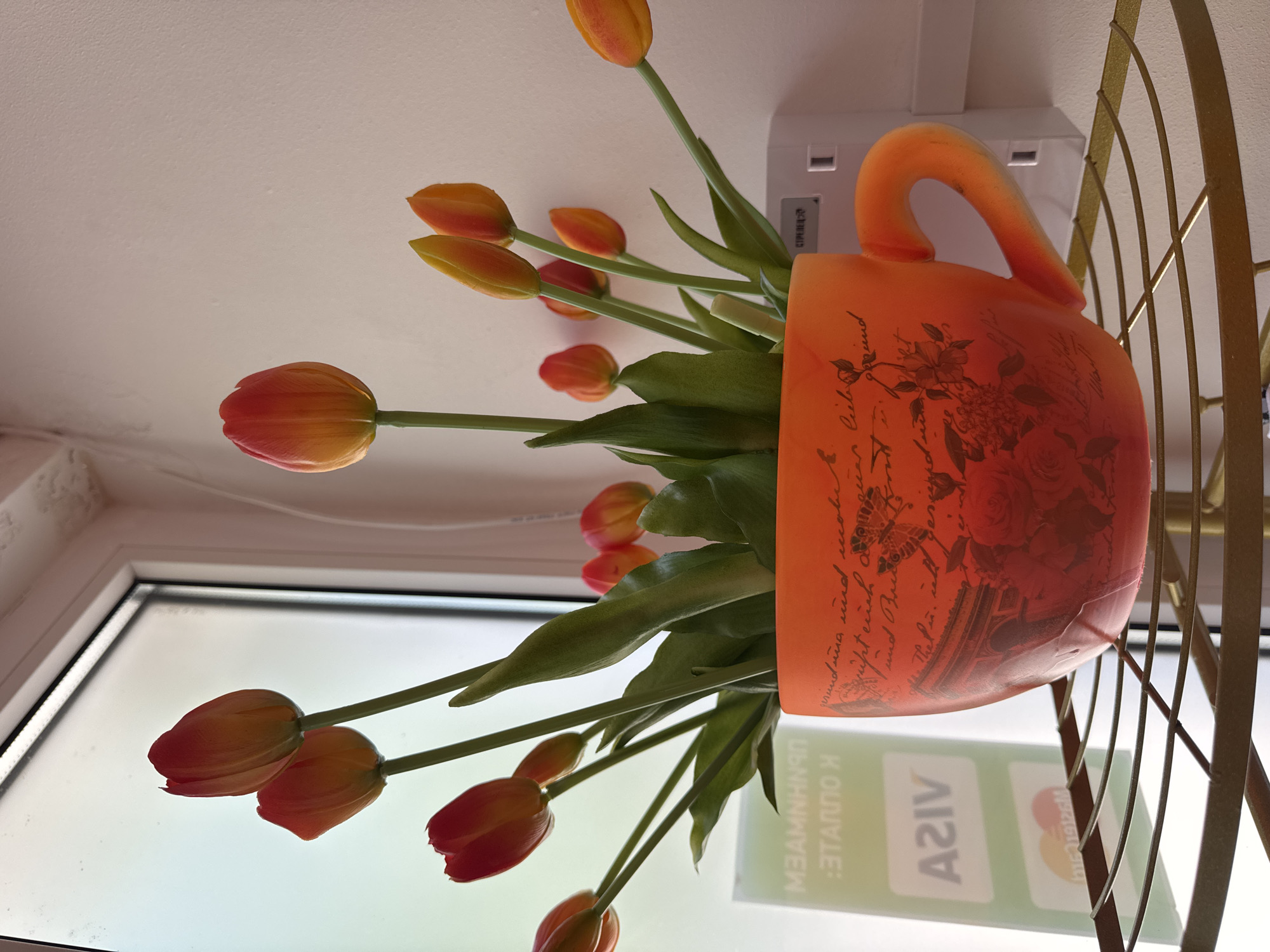 The image presents an upward-looking perspective, featuring a vibrant arrangement of tulips in an ornate orange mug. The mug, positioned on the right side of the frame, has a frosted texture and a prominent curved handle. It is adorned with script-like text, likely German, featuring words such as "Liebe" (love), "und mich" (and me), and "und Dies" (and this), alongside floral and butterfly illustrations.
A bouquet of artificial tulips, showcasing a gradient of orange, red, and yellow hues, emerges from the mug. Their long green stems extend upwards, with most blossoms appearing as closed or partially open buds.
To the left, a white-framed window runs vertically, allowing ample bright, diffused daylight into the scene. Reflections on the window glass include the logos for "VISA" and "Mastercard," along with several lines of reversed Cyrillic text, suggesting an external sign. On the far right, a golden, decorative metal railing with curved bars is partially visible. In the upper-central area, a white rectangular object, possibly a part of a light fixture or sensor, is mounted on a plain white ceiling or wall.
The scene depicts an indoor setting, likely during the daytime, consistent with a shop or cafe in Sochi, Russia, given the visible Cyrillic text and location context. The image captures a static display rather than an active event.