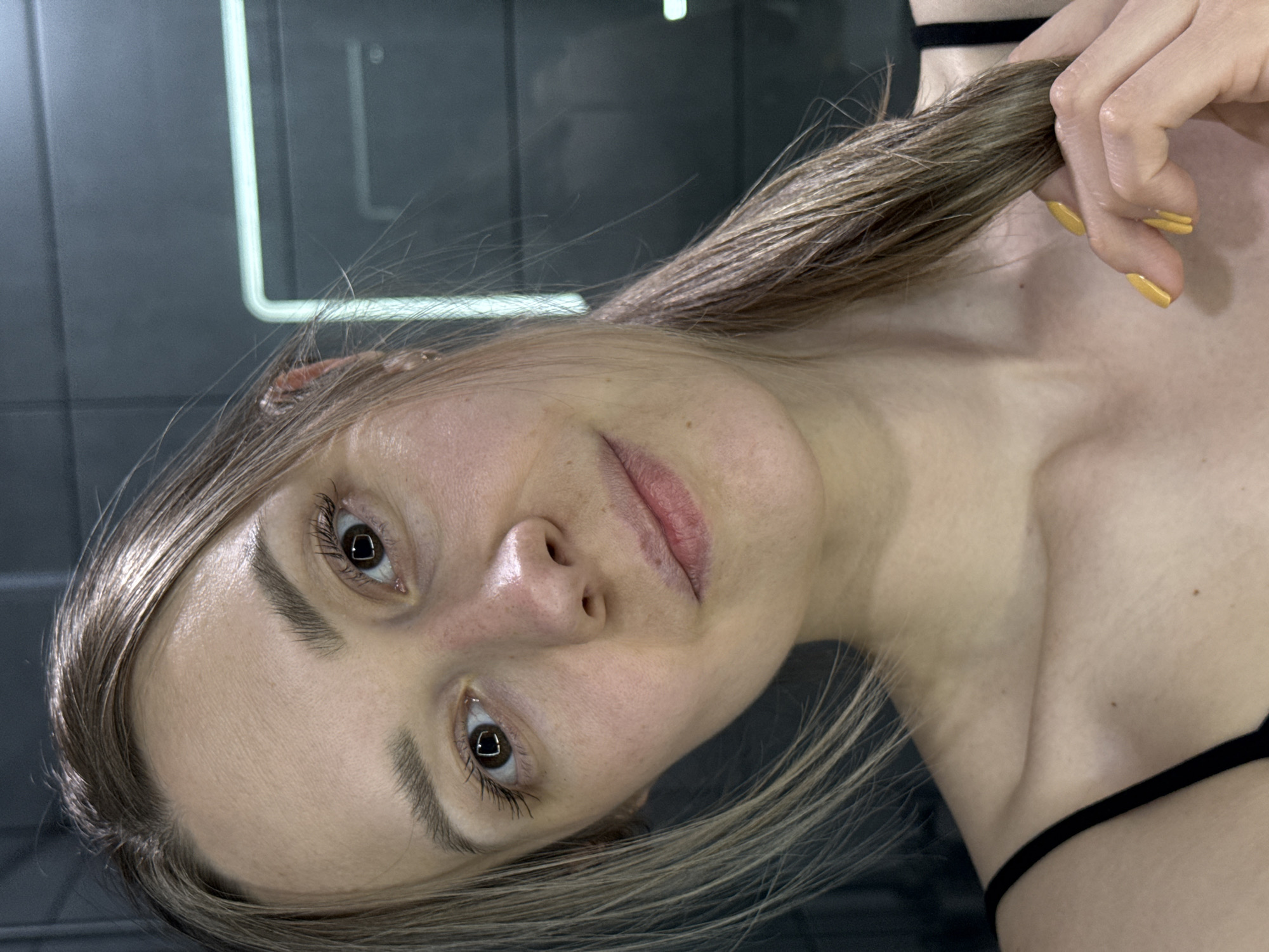 A fair-skinned woman with light brown hair, holding a strand of her hair in her right hand with yellow nail polish, is posing indoors. She is wearing a thin black strap top. The background consists of a dark grey surface with a rectangular light fixture. The city of Chaikovsky, Russia, cannot be confirmed as the location of this image.