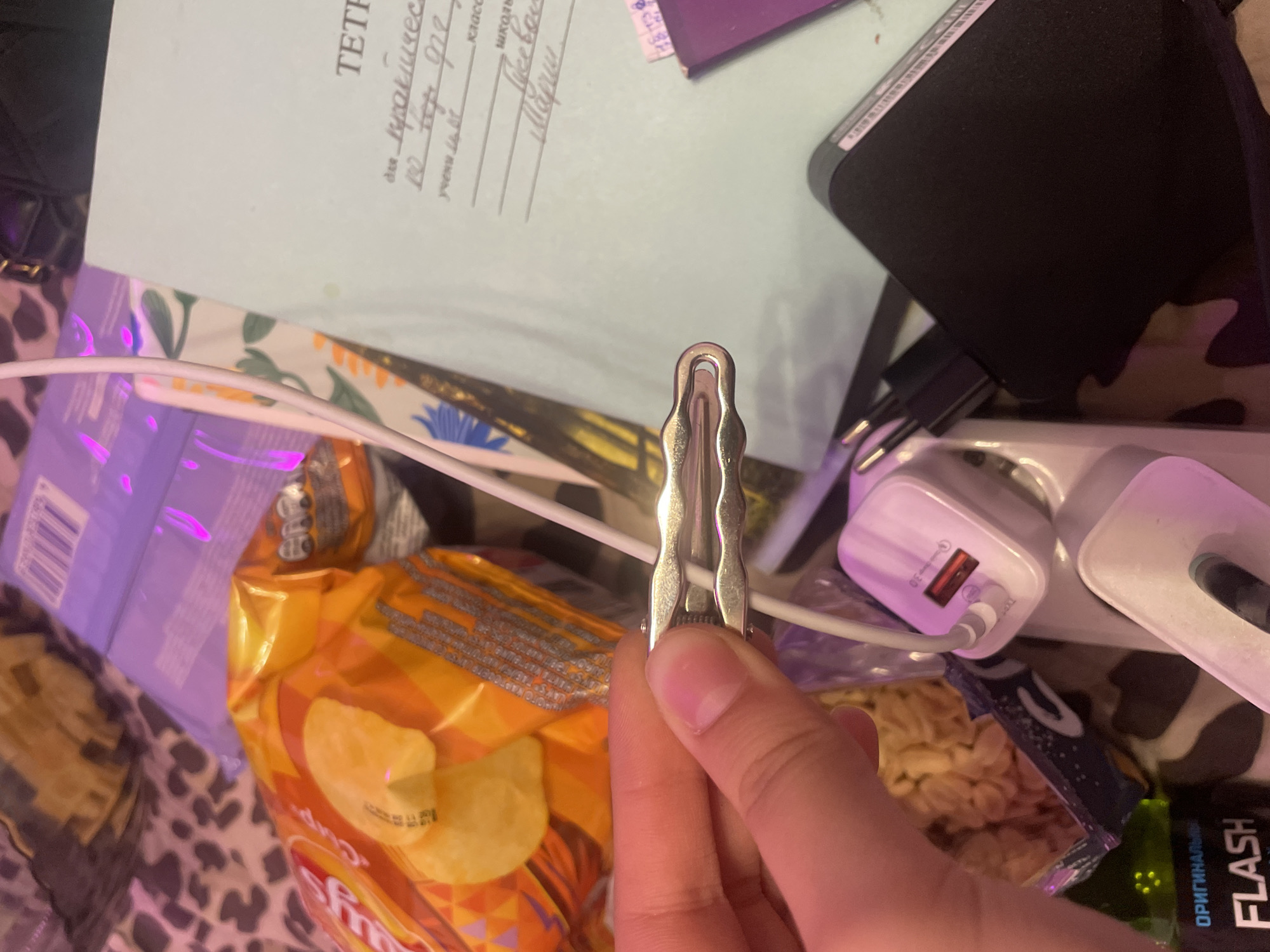 The image displays a close-up view of a person's hand holding a silver hair clip, obscuring a white charging cable and a white charging adapter. In the background, a bag of potato chips, a notebook with handwritten text in Russian, and other electronic accessories are visible. The environment appears to be indoors, with artificial lighting casting a purple hue. There is no indication of the city of Antonovka, Russia, or any other specific location.
