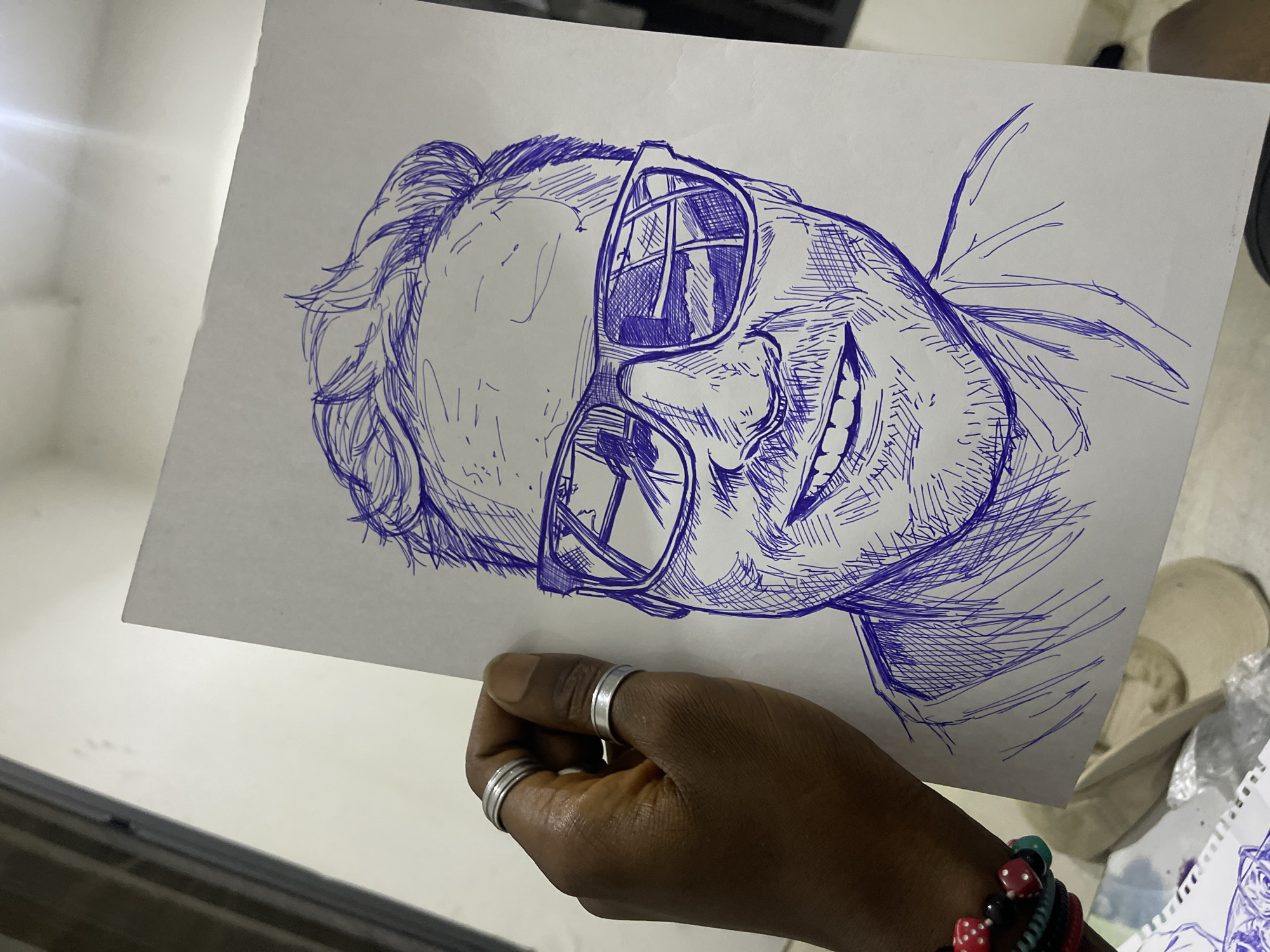 A hand, adorned with two silver rings on the index and middle fingers, holds a blue ink sketch of a man wearing sunglasses and smiling. The drawing is on a white sheet of paper. The background suggests an indoor setting, possibly a room in Abuja, Nigeria, with a wall and a bright light source visible at the top. Other papers, a light-colored cap, and a bracelet made of red and black beads are partially visible in the lower portion of the image.