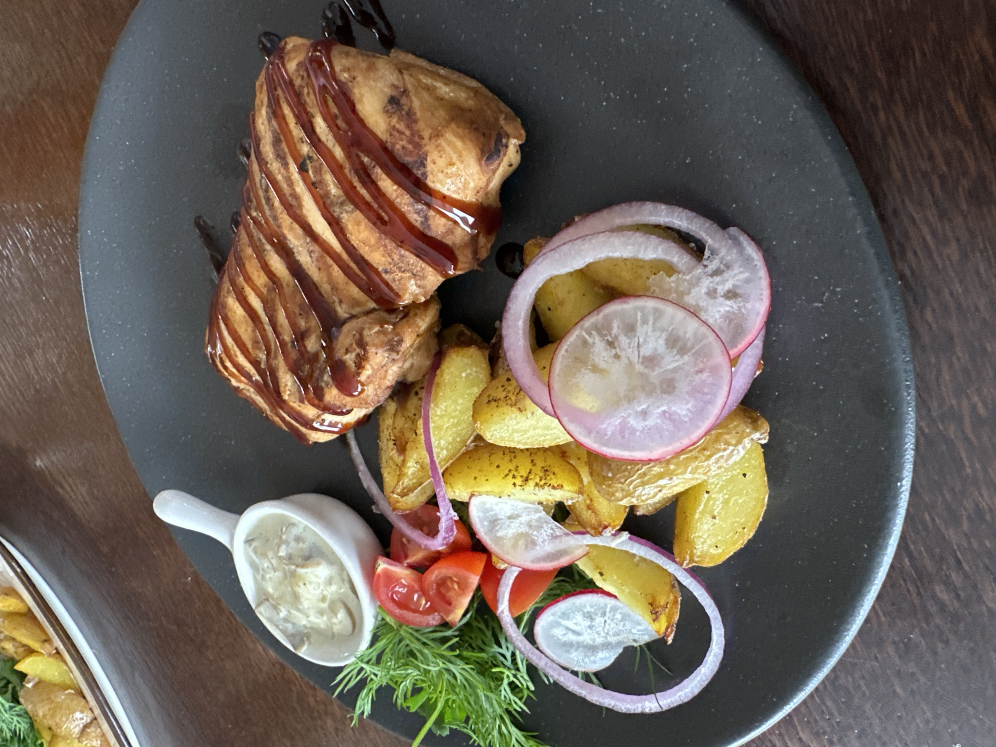 The image shows a plate of food presented on a dark, possibly slate or matte ceramic, oval-shaped plate. The main element on the plate is a piece of cooked, glazed meat, drizzled with a dark sauce, resembling a barbecue or teriyaki glaze. To the right of the meat, there's a serving of golden-brown roasted potatoes, garnished with sliced red onion and thinly sliced radishes. A small scoop of white sauce, likely a mushroom or cream-based sauce, sits in a small white dish. Additionally, there are tomato slices and sprigs of fresh dill. The plate rests on a dark wooden surface.
Based on the image context provided (Chaikovsky, Russia), it could be a dish served in a restaurant or prepared in a home in that location.