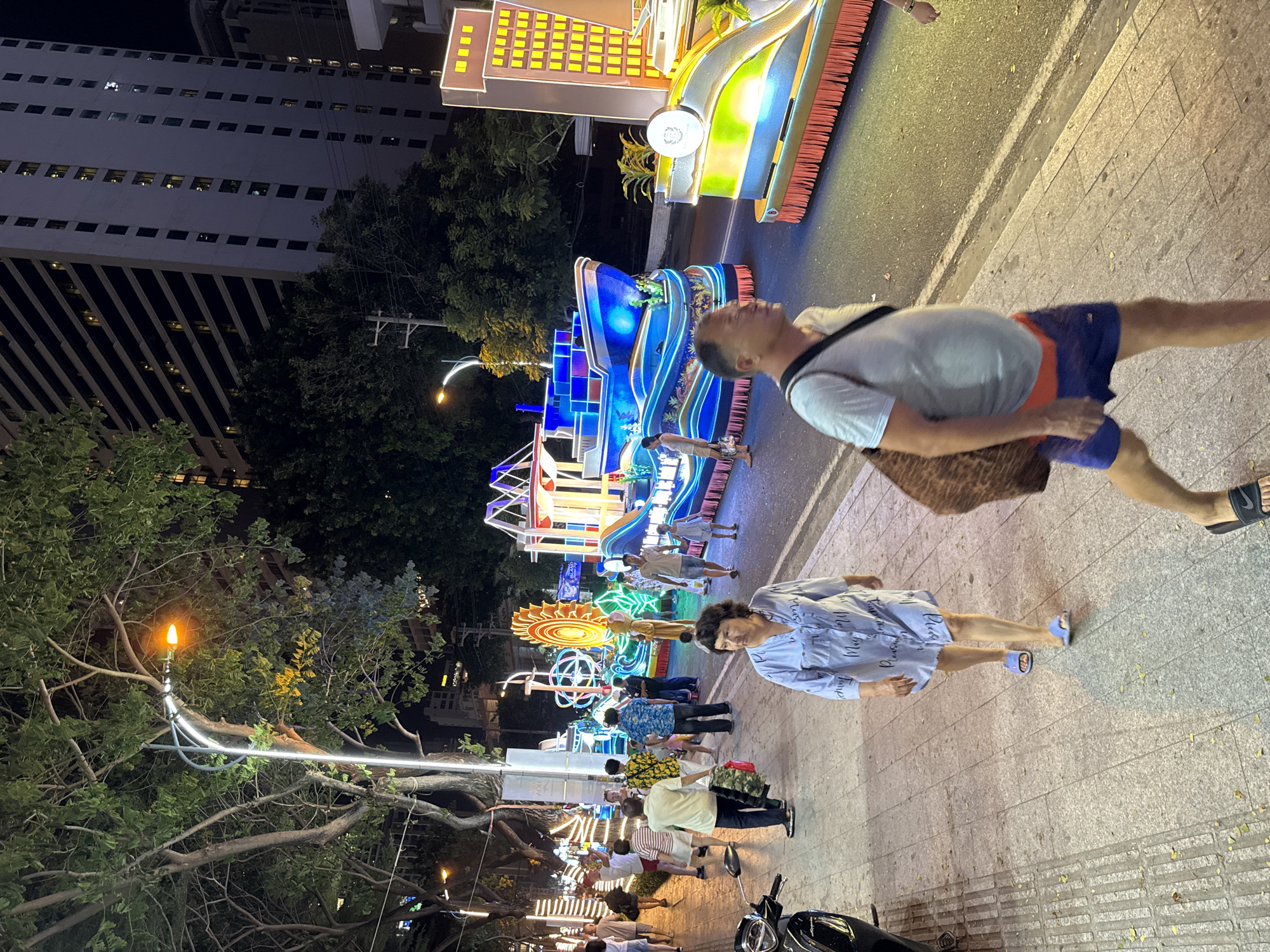 In Nha Trang, Vietnam, at night, a street is illuminated by decorative lights and floats. A parade is in progress, featuring a large blue float shaped like a ship, adorned with lights and depicting an underwater scene. Another float, resembling a building, is also present. Several people are walking on the sidewalk and observing the parade. A woman in a light blue dress walks towards the viewer, and a man in a light blue t-shirt and swim trunks walks past her, carrying a bag. A motorcycle is parked on the left side of the image. The street is paved, and trees line the sidewalk. Buildings are visible in the background.