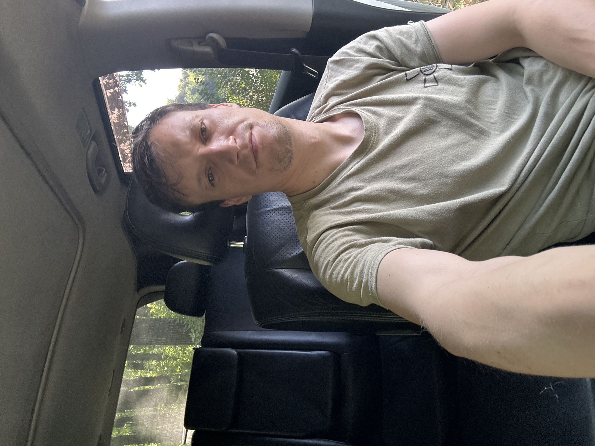 A man is sitting in the back seat of a car, taking a selfie. He is wearing a light brown t-shirt with a black symbol on the left chest. The interior of the car is visible, including black leather seats and the car's headliner. Through the windows, a wooded area with green foliage is visible, suggesting a rural or suburban environment. The lighting indicates it is daytime. The man is looking directly at the camera with a neutral expression. His right arm is extended, holding the phone for the selfie. The image captures the man from the chest up, with the background of the car interior and the outdoor scenery visible.