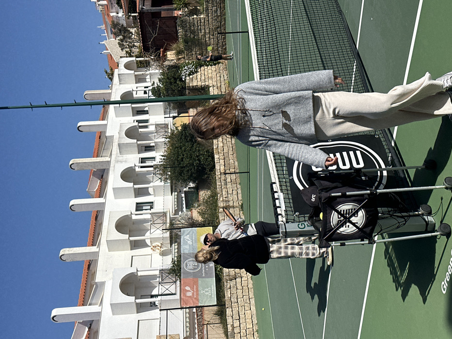 The image depicts a bright, sunny outdoor scene on a green tennis court in Ericeira, Portugal. In the foreground, positioned to the right of a black tennis net with a white top band, stands a female with long, wavy brown hair. She wears a grey knit hooded sweater, light beige wide-leg ribbed pants, and light-colored sneakers. Her face is partially obscured by her hair, and sunglasses hang from the front of her sweater. Adjacent to the net is a black wheeled ball cart containing yellow tennis balls, a black cap with "arc" text, and a black bag with a white circular logo featuring intersecting lines. A larger black banner with white vertical lettering and a circular logo is positioned behind the cart.
In the midground, to the left of the net, a blonde female wearing a black top and plaid pants stands on a dark, flat board, possibly a skateboard. She is facing towards the center-right. Behind her, a male wearing a light-colored baseball cap, grey hoodie, and dark pants holds a brown, wooden-appearing tennis paddle or training racket. A yellow tennis ball lies on the court surface near the blonde female.
In the far background, beyond a green wire fence and a stacked stone wall, a male figure is visible, engaged in playing tennis on another part of the court or an adjacent court. He wears a dark shirt and shorts and holds a tennis racket.
The background is dominated by a row of white residential buildings with terracotta tiled roofs, multiple arched balconies, and distinctive tall, curved chimneys, characteristic of Portuguese architecture. Vegetation, including bushes and small trees, is visible along the stone wall and in front of the buildings. A banner with "tennis school" and other logos is visible on the fence. The sky is clear and blue, and the scene is illuminated by strong sunlight, casting visible shadows.