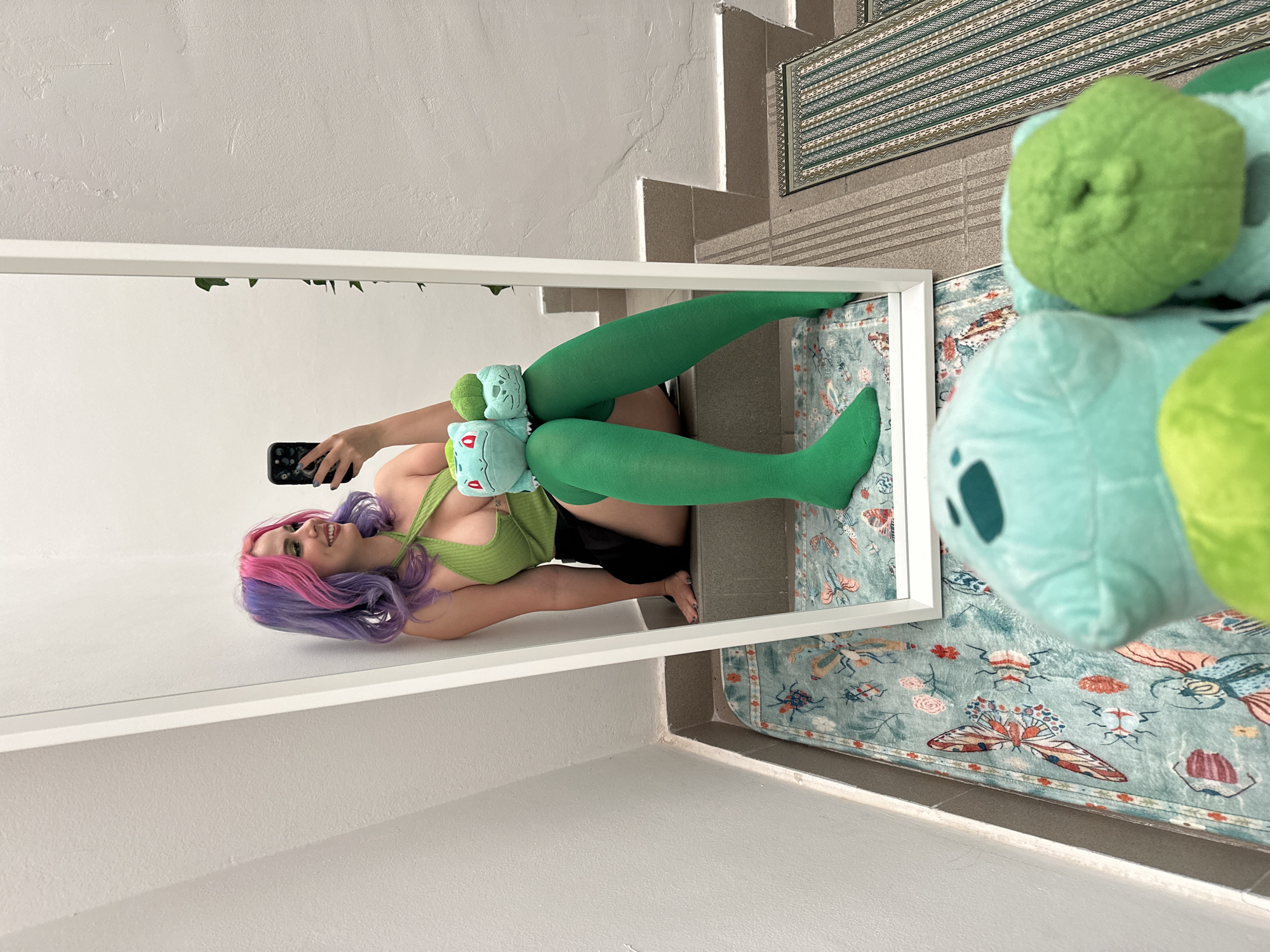 A young woman is posing for a mirror selfie. She has purple and pink hair, wears green thigh-high stockings, a matching green top, and black shorts. She is holding two Bulbasaur plush toys. The mirror frame is white. The background includes a light-colored wall, a staircase, a patterned rug, and a large Bulbasaur plush toy partially obscuring the view. The picture was taken in City of Zagreb, Croatia.