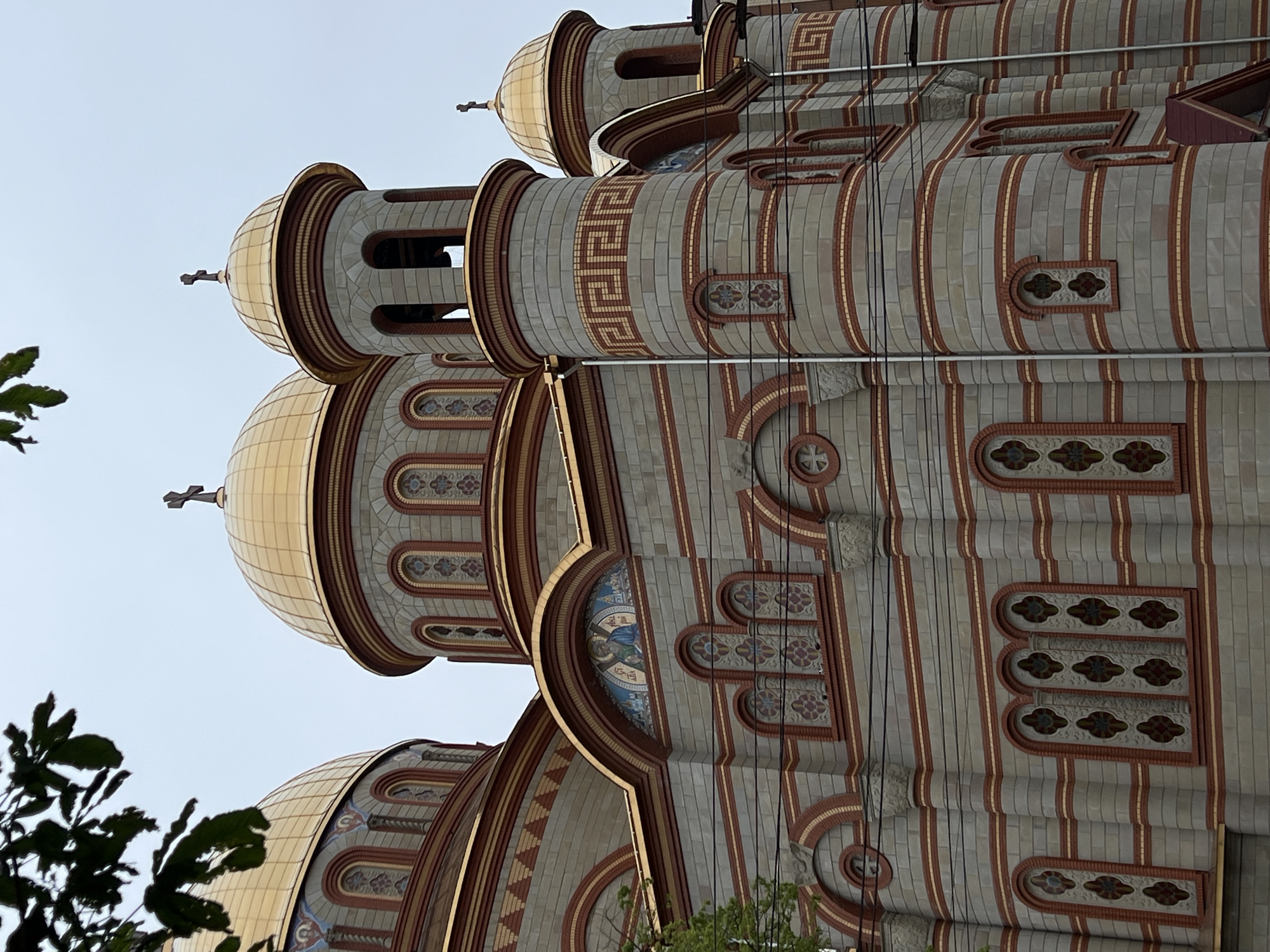 The image presents a close-up view of an elaborate Eastern Orthodox church, seen from a low angle looking upwards, characteristic of structures found in places like Ternopil, Ukraine. The church is distinguished by its multiple prominent golden, onion-shaped domes, each topped with a cross, reflecting light under a clear, bright sky.
The facade of the church is a tapestry of intricate design, featuring alternating bands of light grey and reddish-brown brickwork. These bands are arranged horizontally and vertically, creating a textured surface. Geometric patterns, notably Greek key (meander) motifs, are intricately integrated into the cylindrical elements supporting the domes, and along other sections of the building. Numerous arched windows and recessed panels adorn the walls, some containing colorful mosaic artwork depicting religious figures, such as a figure in blue within one arch, while others display decorative, stained-glass-like patterns or ornamental brickwork designs.
On the left side of the frame and towards the bottom, green foliage from nearby trees partially obscures the church, offering a natural element against the constructed grandeur. Several black wires run vertically across the facade of the building, possibly utility lines. The clear sky indicates a bright, daytime setting. The opulent golden domes and detailed facade are typical architectural features of Orthodox churches. No visible text is present in the image.