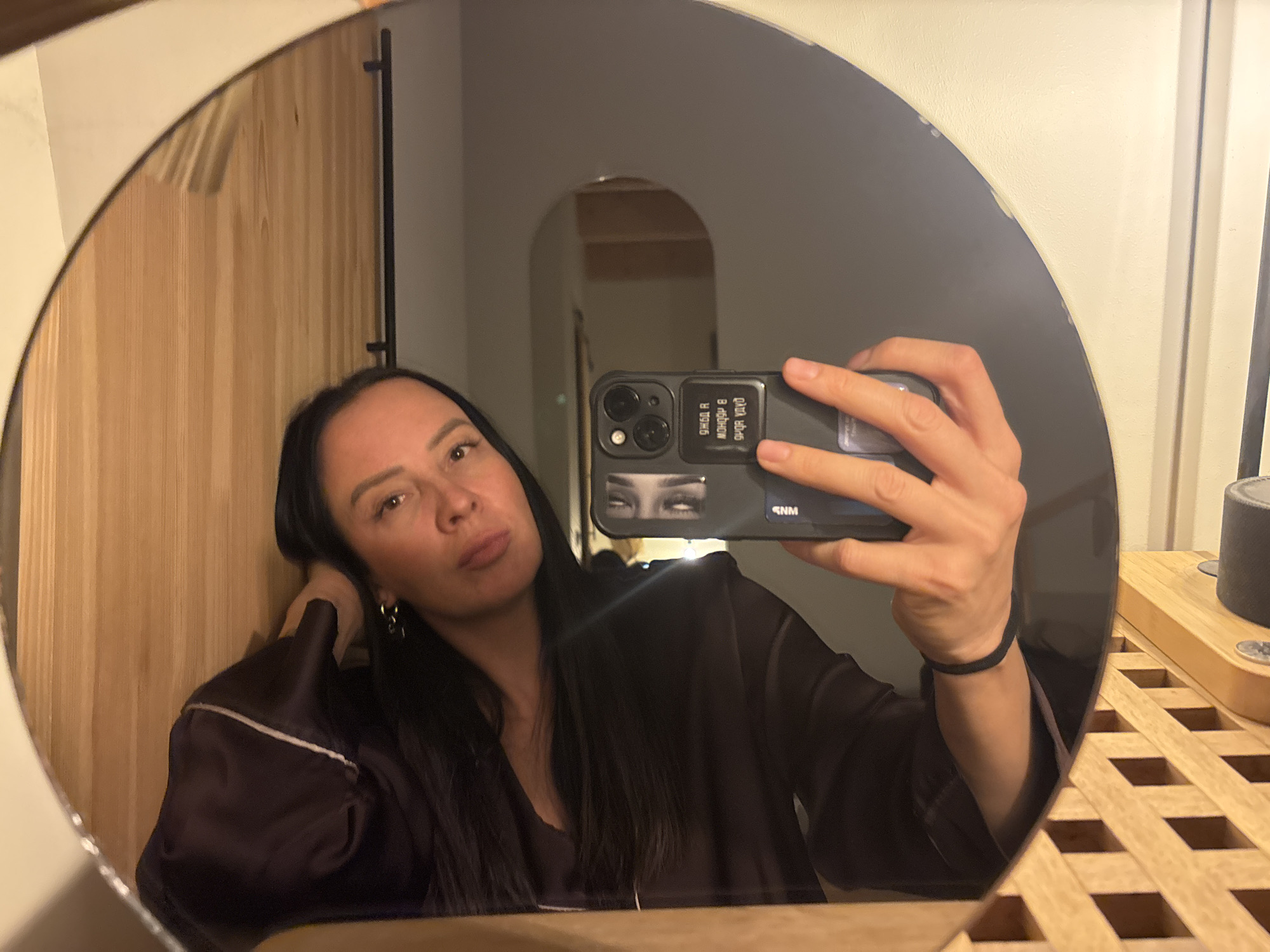 A woman is taking a selfie in a round mirror. The background shows an interior space with light wood paneling on the left and a doorway leading to another room straight ahead. A dresser or shelf with a latticework pattern is visible on the right. The phone case appears to have a graphic of eyes and reversed text. The woman is wearing a dark top with light trim. The location is in Chaikovsky, Russia.