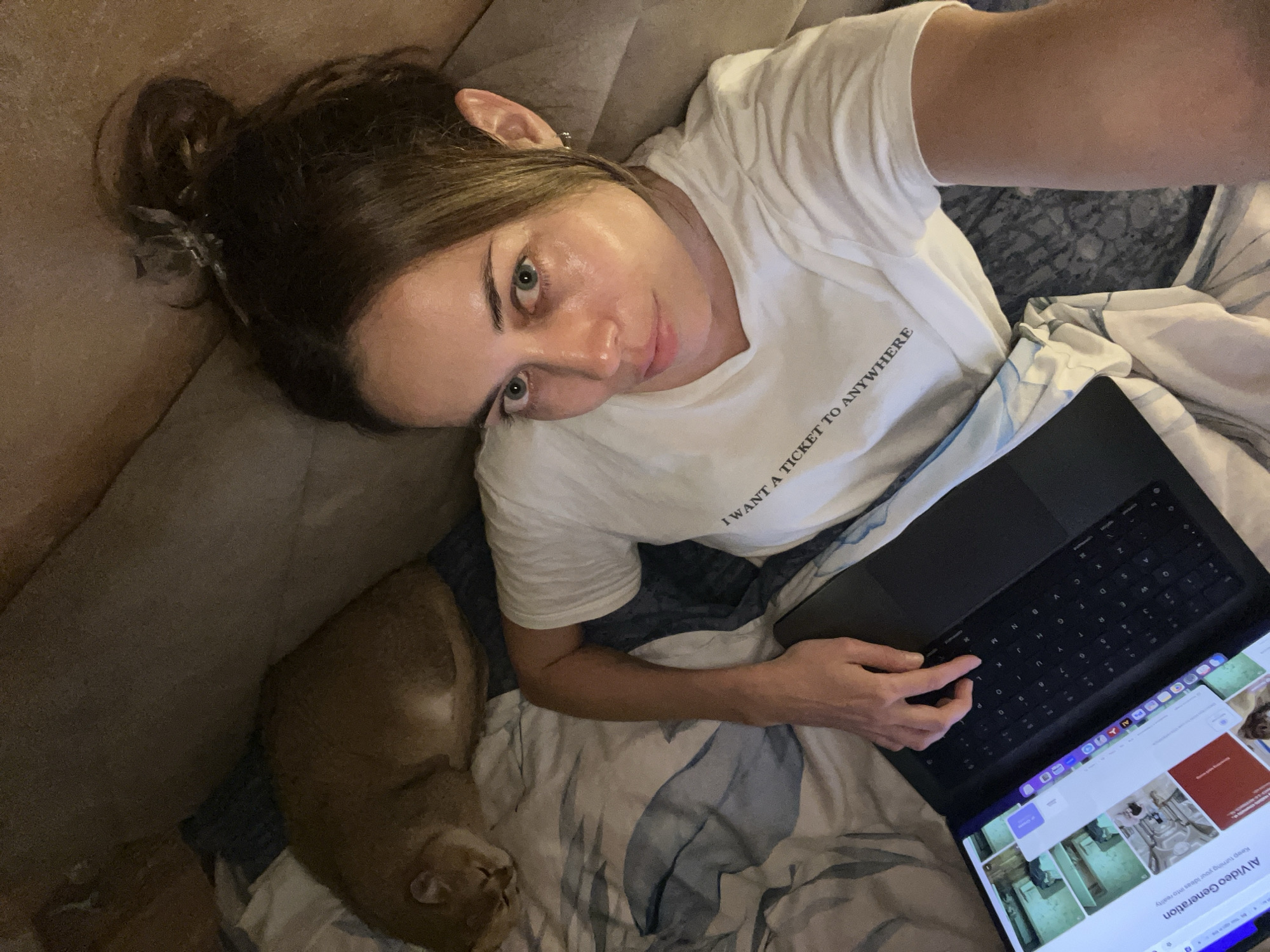 A woman lies on a bed with blue and white sheets, holding a black laptop displaying "AI Video Generation" on the screen. She is wearing a white t-shirt with the text "I WANT A TICKET TO ANYWHERE". Her hair is pulled back in a bun, and she is looking at the camera. A tan-colored cat is lying to her left. The scene is indoors, possibly a bedroom. The location in the city of Bar, Montenegro is not apparent from the image.