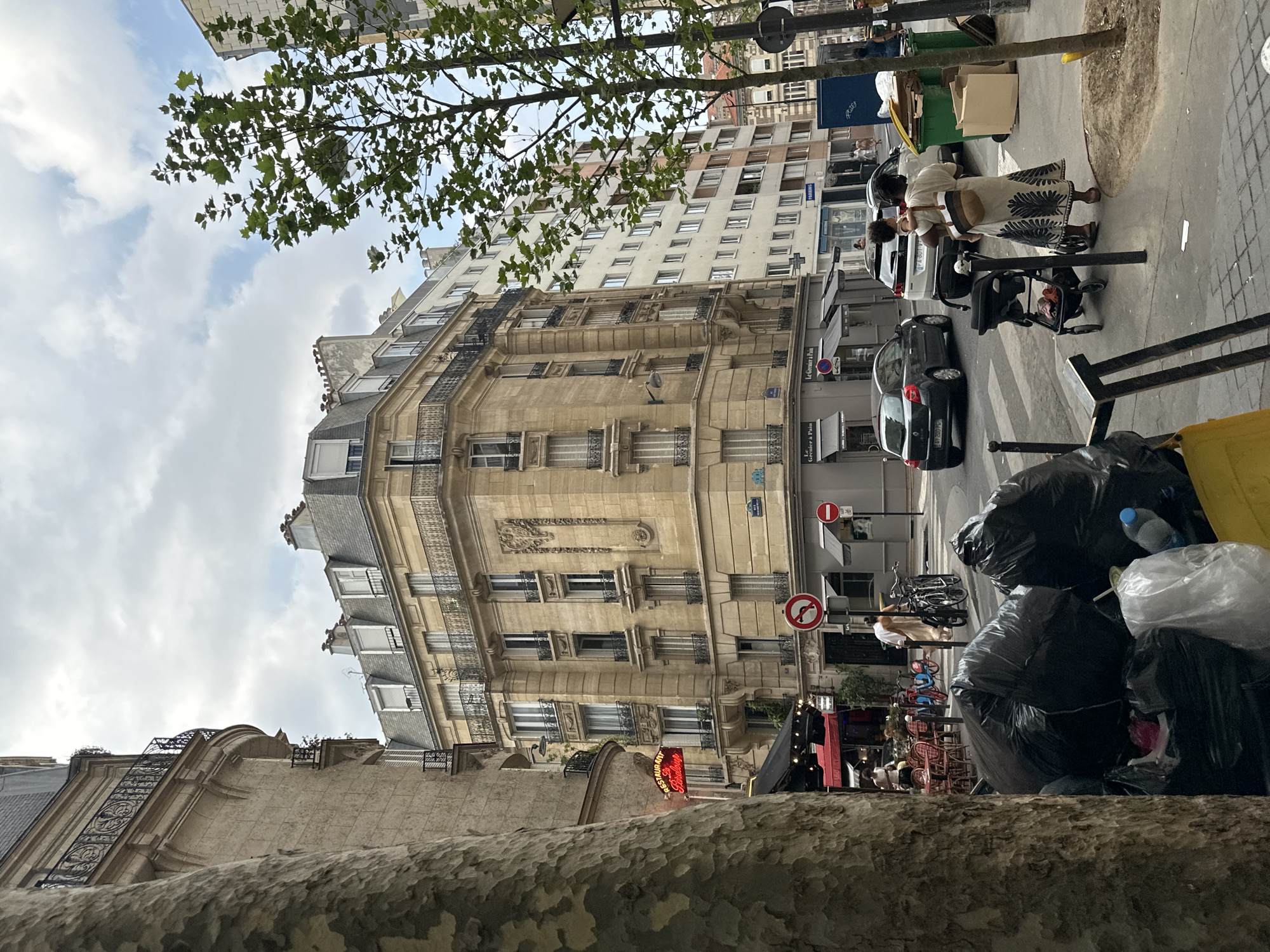 The image depicts a street scene in Paris, France, under a cloudy sky. In the foreground, several large black garbage bags are piled up on the sidewalk, partially obscuring a yellow bin. A large tree trunk is on the far left. Beyond the garbage bags, a street intersection is visible.
On the left side of the street, a building with a red neon sign that reads "RESTAURANT La Perle" is situated next to an outdoor cafe with red chairs and tables. Further down the street, a corner building with ornate beige stone facade and balconies stands prominently. To the right of this building, a taller, modern white building is visible.
Several cars are on the street, including a dark grey Renault Clio and a white Peugeot SUV. People are also present: a woman is carrying a child on her hip and walking with a stroller, while other pedestrians are seen further down the sidewalk. Bicycles are parked near the cafe. Traffic signs, including a "no left turn" sign, are visible at the intersection. Green and brown recycling bins are also present on the sidewalk to the right of the street. The overall scene suggests a bustling urban environment with a mix of classic and modern architecture.