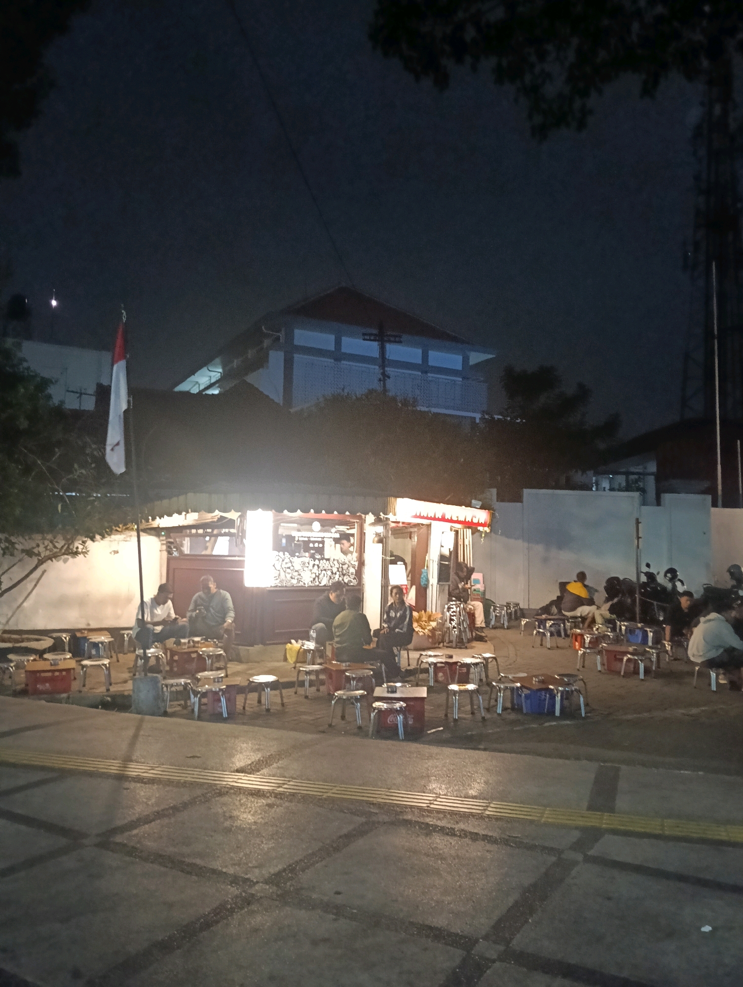 The image depicts a street scene at night in Malang, Indonesia, likely near a popular gathering spot. In the foreground, a paved sidewalk with tactile paving for the visually impaired is visible, showing tire tracks and reflective surfaces from lighting. Beyond the sidewalk, a cluster of small tables and stools are arranged outdoors, many with red or blue containers on them.
A food stall with bright overhead lighting and a red and white awning is the central focus. Several people are seated at tables and standing around the stall, engaged in conversation or ordering food. A flag, appearing to be the Indonesian flag with red and white colors, is displayed on a pole to the left of the stall.
In the background, there are buildings, including one with multiple levels and balconies. A tall telecommunication tower is visible on the right side of the image. The overall atmosphere suggests a lively, informal nighttime market or a roadside eatery popular with locals. The lighting creates a warm, inviting glow around the stall and the seating area.