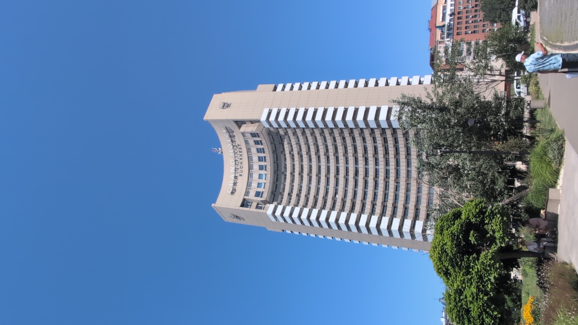 The image shows the InterContinental Hotel in Bucharest, Romania, a tall, light-brown building with a curved facade and white horizontal bands. The sky is clear and blue. In the foreground, there is a park with trees, benches, and people. Two individuals are seated on a bench under a large, leafy tree. Further to the right, a person wearing a white cap and a patterned shirt is walking on a paved path, holding a mobile phone. In the background, other buildings are visible. The overall scene depicts a sunny day in a city park adjacent to a prominent hotel.