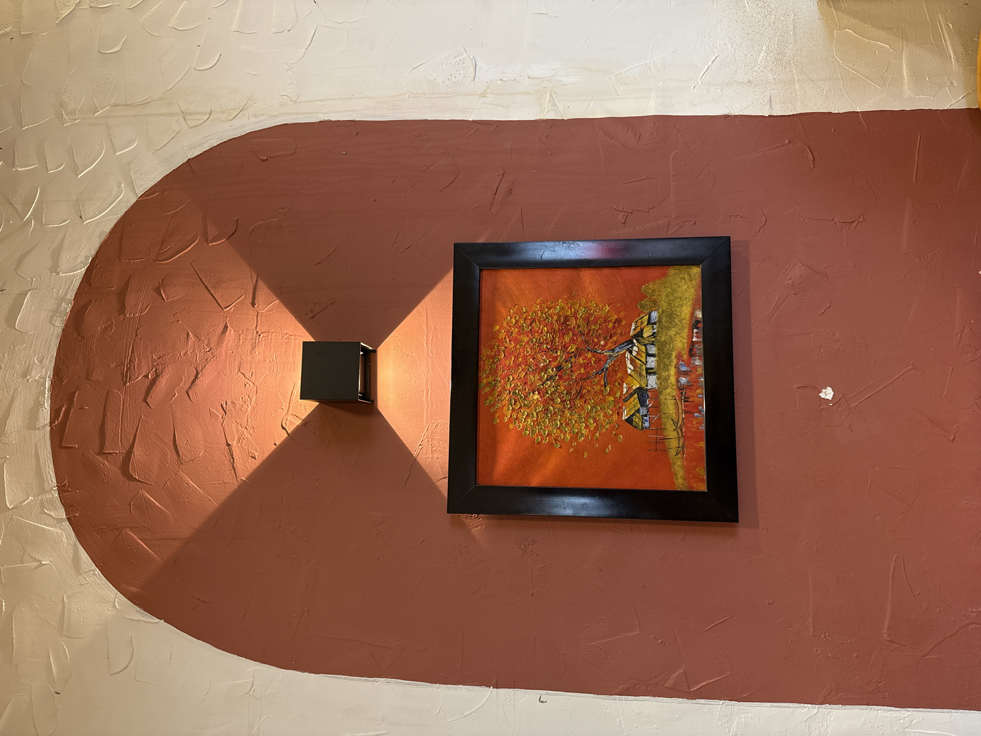 The image shows a section of a textured wall in what appears to be an interior space, likely a restaurant or shop. The wall is divided into two distinct sections: the lower section is painted in a light, off-white color, while the upper section is painted in a rust-brown or terracotta hue. These two sections are separated by an arch-shaped border.
Mounted on the rust-brown section of the wall is a framed painting depicting a tree with orange and yellow foliage, possibly an autumn scene. The frame is black. To the left of the painting is a modern, black cube-shaped sconce, emitting a beam of light that casts a triangular shadow across the wall. A small, white object is stuck to the wall to the right of the painting.
The textured effect of the wall is created by what appears to be a textured paint or plaster application.
Based on the request, the location is in Bac Nha Trang Ward, Vietnam.