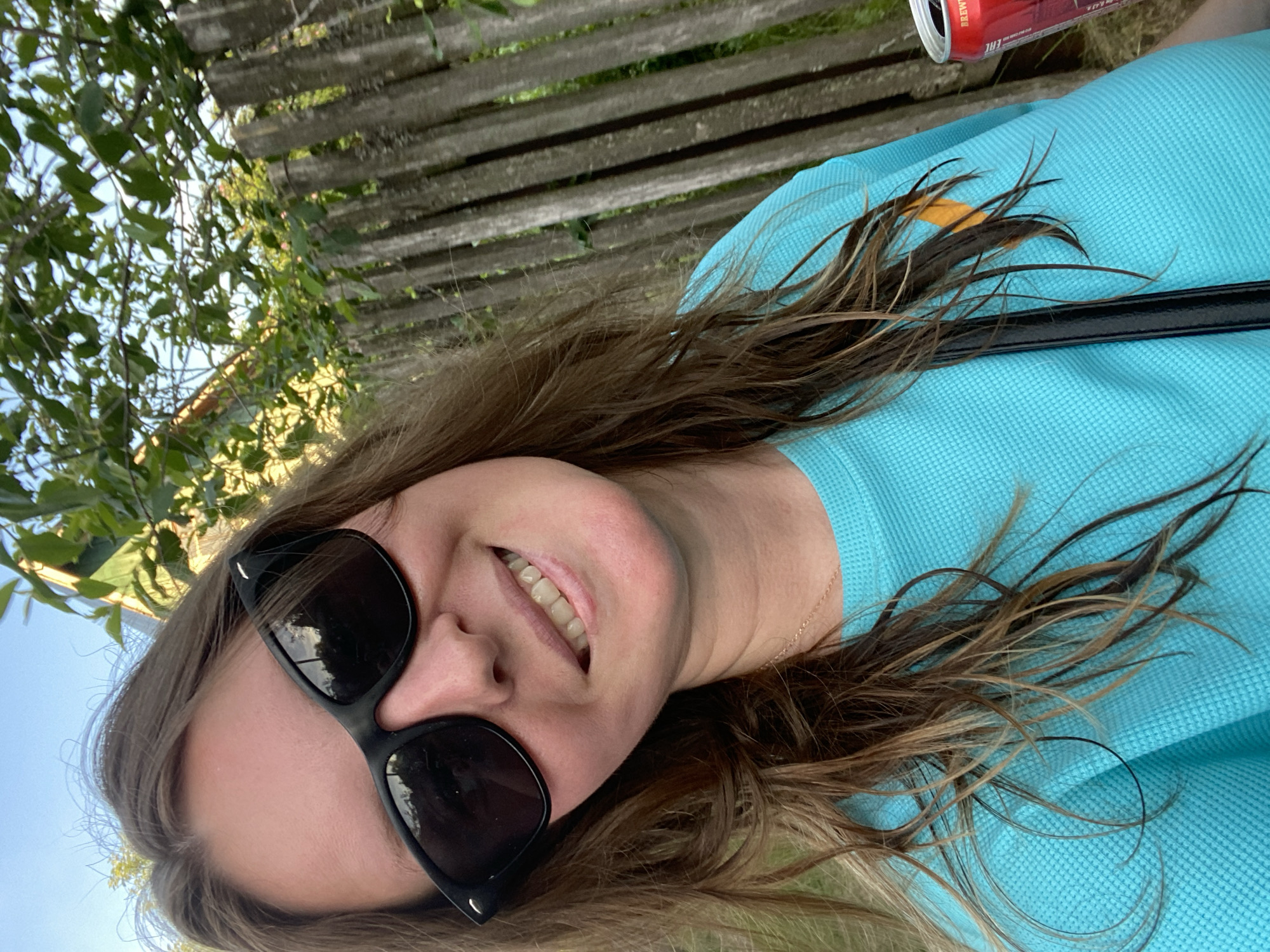 A close-up, tilted outdoor portrait features a smiling woman with long, wavy brown hair and blonde highlights. She wears large black sunglasses that cover her eyes and a light blue (turquoise) waffle-knit polo shirt, with a thin gold chain necklace visible. Her wide smile reveals her teeth, conveying a joyful and relaxed emotion.
The background shows a weathered wooden fence constructed from horizontal planks. Lush green foliage and trees are visible behind and above the fence, suggesting a natural, possibly park-like or garden setting. A bright blue sky is visible in the upper left, indicating clear, sunny daytime weather.
In the upper right, a red aluminum beverage can is partially visible near the woman's shoulder and hair. Faint white text on the can includes "BREW", "EAC", and numbers like "10". A black strap, likely from a bag, also rests on her shirt. The image captures a happy, casual moment outdoors, with the context being Chaikovsky, Russia.