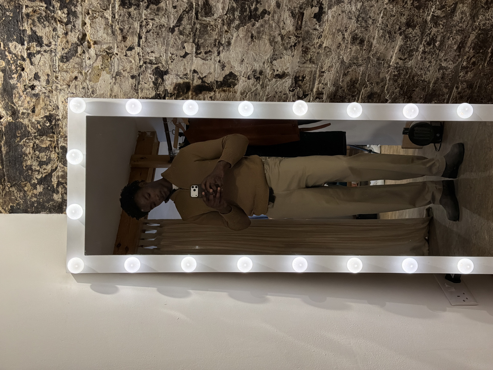 A full-length mirror with light bulbs around the perimeter reflects a Black man standing in front of it. The man is wearing a light brown sweater, tan pants, and gray shoes. He is holding a cell phone. Behind the man and reflected in the mirror is a wooden structure and various articles of clothing hanging. The wall behind the mirror is made of rough stone or brick, while the wall in front of the mirror is a light color. An electrical outlet is visible on the wall below the mirror. The location is potentially in London, United Kingdom.