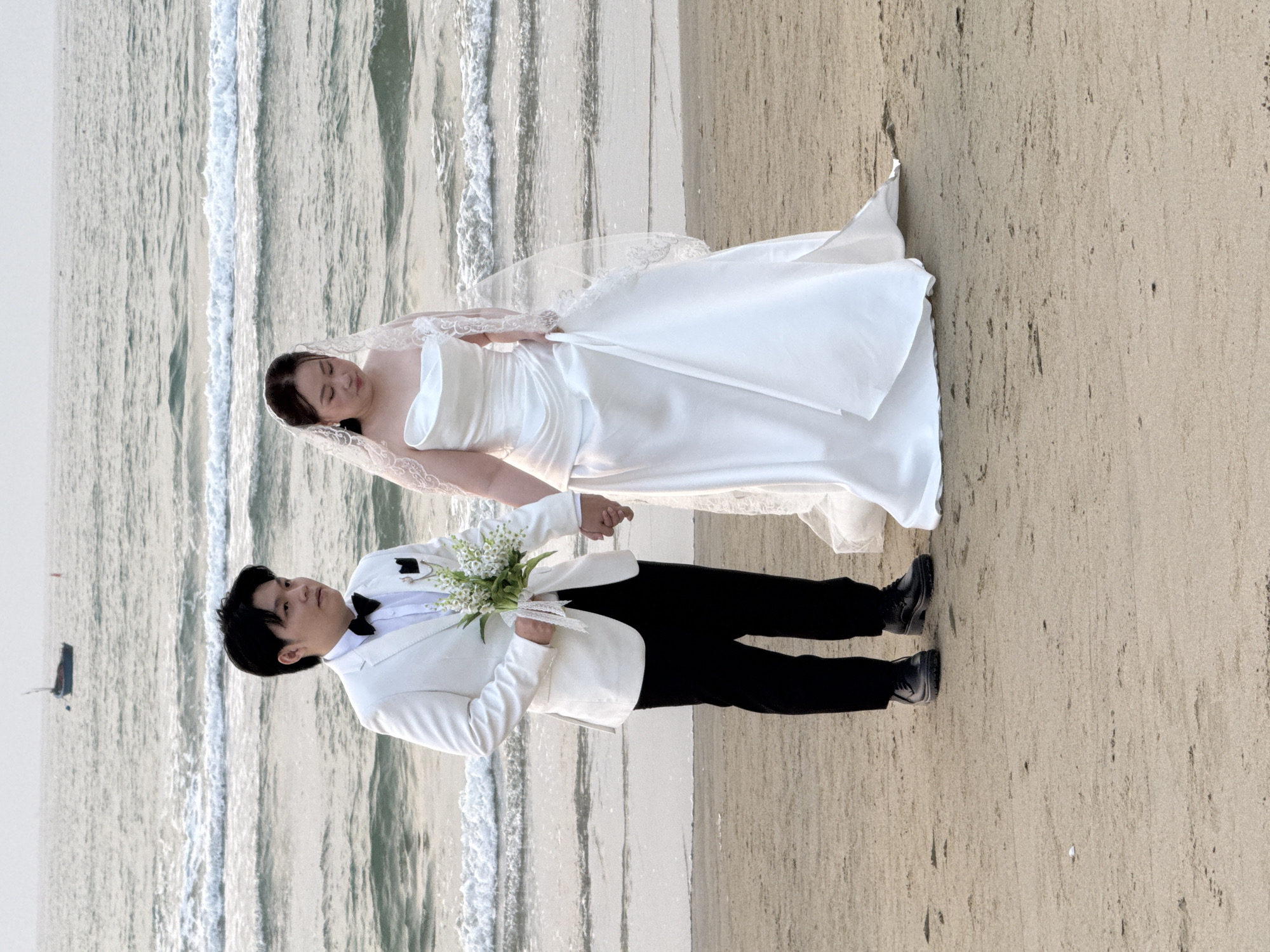 A wedding photo shows a groom and bride holding hands on a beach. The bride wears a white strapless dress and a lace-edged veil. The groom wears a white suit jacket, a black bow tie, and black pants. He holds a bouquet of white flowers. The background includes the ocean with visible waves and a sandy beach. A small boat is visible in the water in the background. The location is Đà Nẵng, Vietnam.