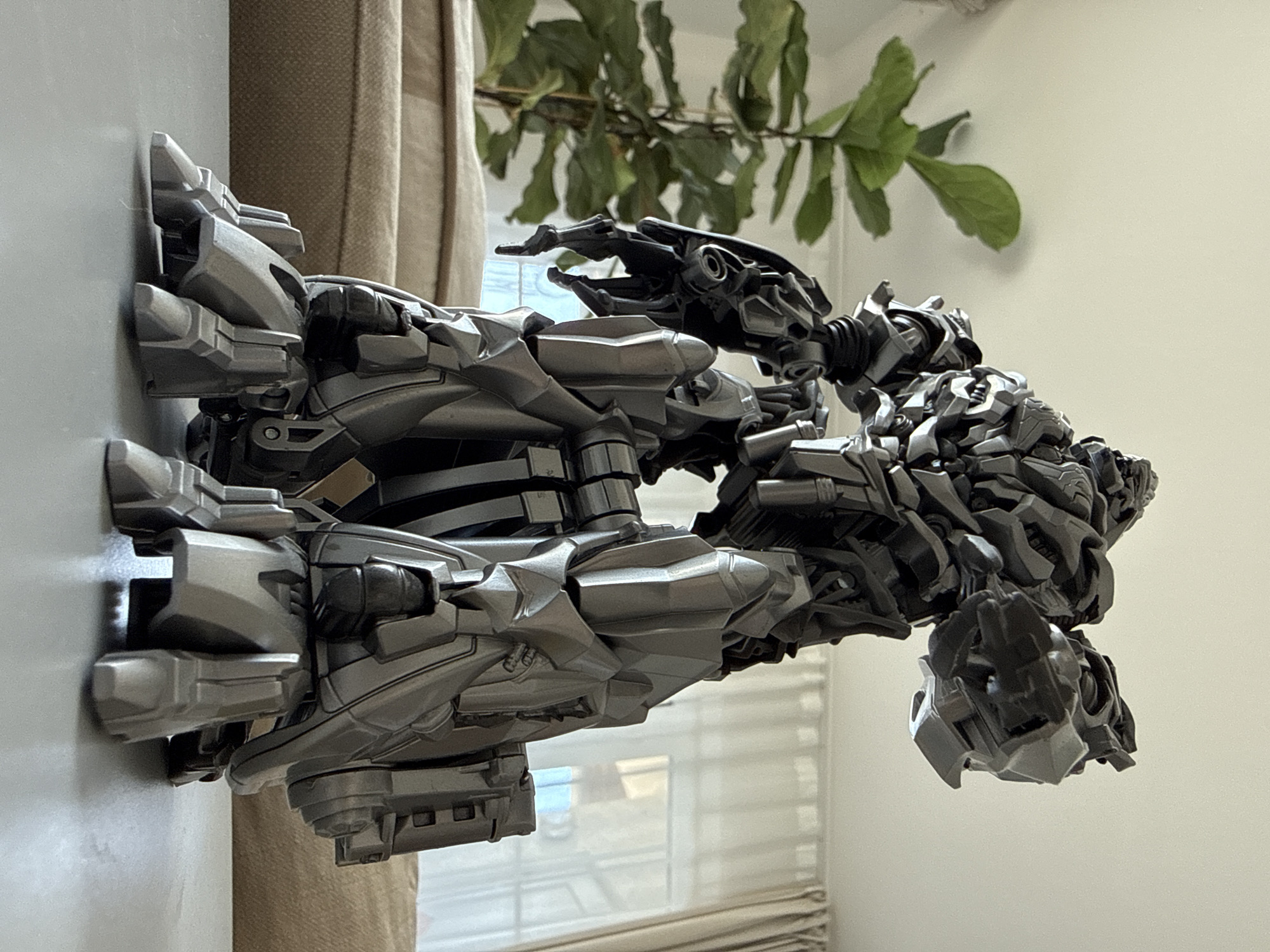 A detailed, grey, robotic action figure, likely Megatron from the Transformers franchise, stands on a grey, flat surface. The figure is highly articulated, with numerous mechanical components visible in its body, arms, and legs. The figure is positioned in a dynamic pose, with its arms extended and its head turned upwards. In the background, a green, leafy plant is visible to the left, and sheer, off-white curtains hang in front of a window to the right. A beige upholstered couch is visible behind the figure. The overall setting appears to be an indoor environment, possibly a living room. There is no indication of the city of Bad Sachsa, Germany, or any specific urban location.