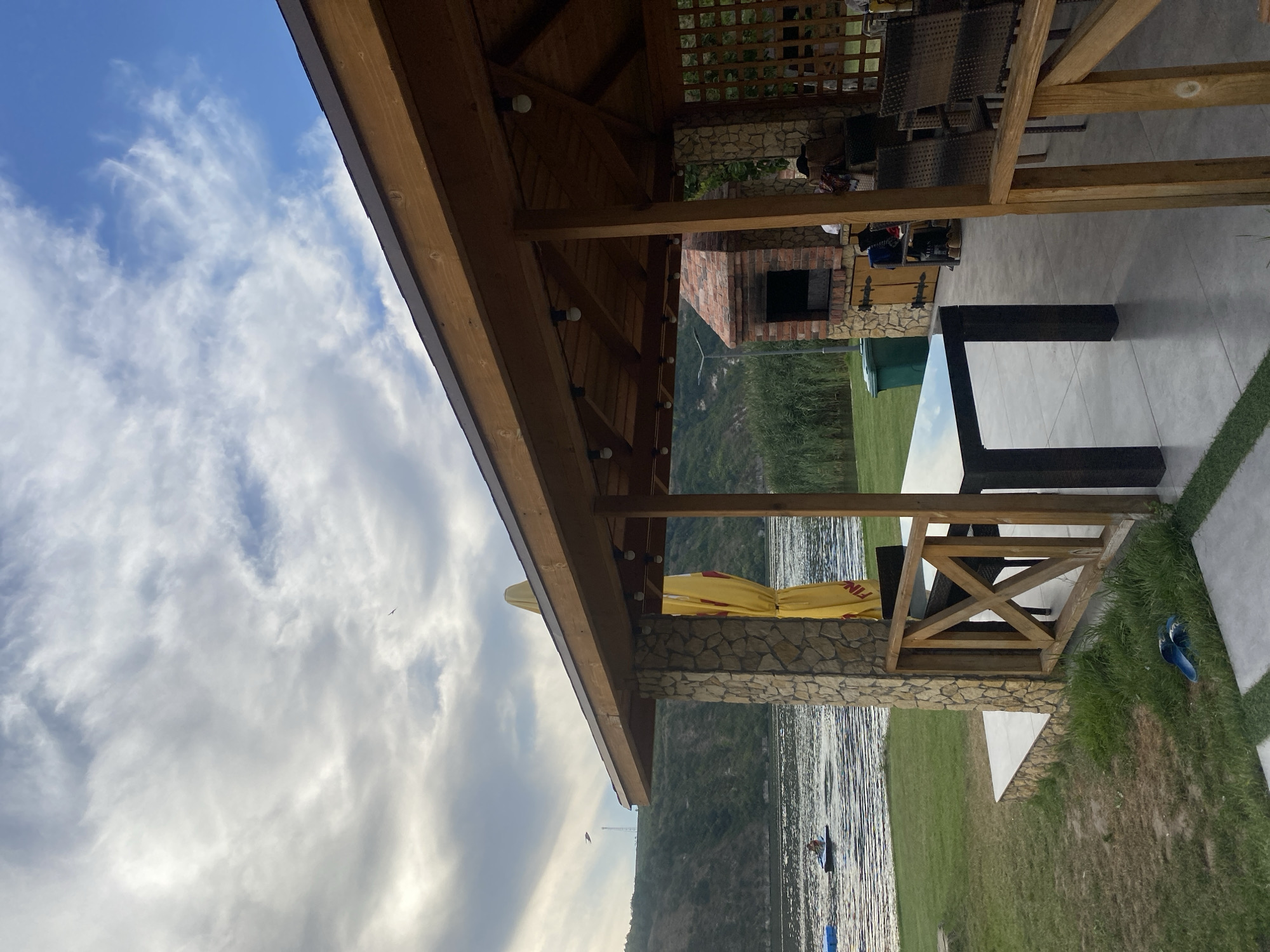 A covered patio overlooks a body of water in Molovata Nouă, Moldova. The patio is constructed of wood with stone pillars and features a table and chairs. A brick barbecue grill is visible behind the seating area. In the distance, a hill covered in vegetation rises from the water's edge. The sky is partly cloudy.