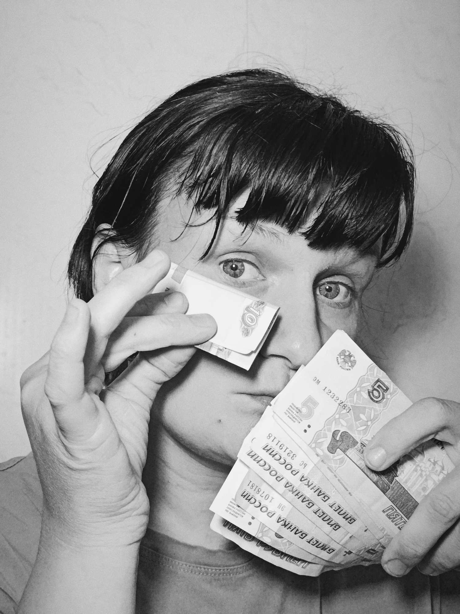 A black and white close-up photograph captures an individual, likely an adult, with short, dark hair and bangs, holding Russian ruble banknotes to their face, partially obscuring it. The person's right eye is visible, wide open, and directed towards the viewer with an intense gaze. Their left hand holds a 10 ruble banknote near the left eye, while their right hand holds a fan of several 5 ruble banknotes over the lower part of their face.
The banknotes are prominently displayed, revealing text such as "БИЛЕТ БАНКА РОССИИ" (Bank of Russia Bill), the denominations '5' and '10', and serial numbers including "ЭМ 1232283", "9H 1078181", and "BC 3219118". Other visible text includes "ПРЕСЛЕДУЕТСЯ ПО ЗАКОНУ" (persecuted by law), likely a warning against counterfeiting. The background is a plain, light-colored wall, indicating an indoor setting. The image, taken in Фоки, Russia, conveys a sense of preoccupation or focus on money.