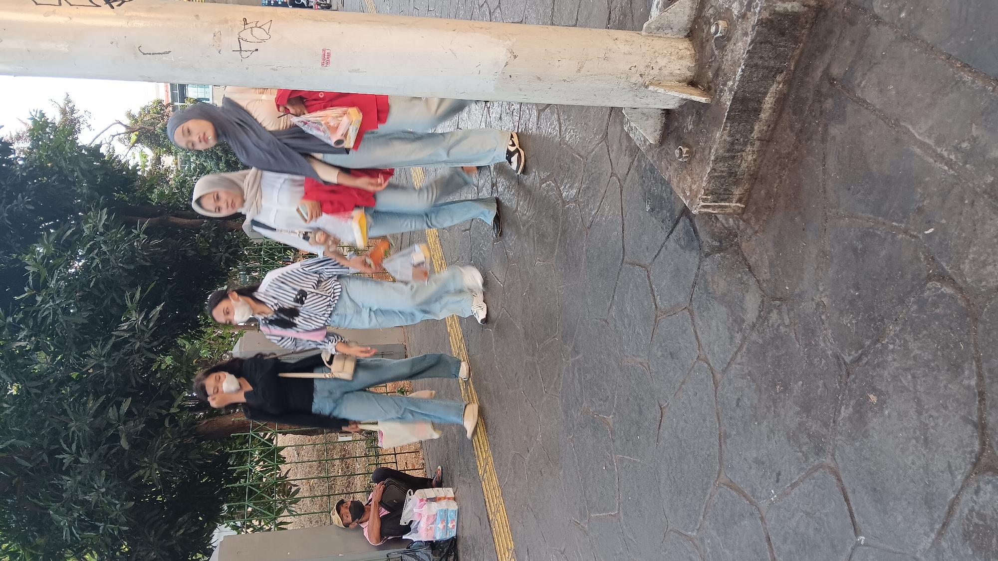 In South Jakarta, Indonesia, a street scene unfolds with four young women walking on a paved sidewalk. Three of the women are wearing face masks, suggesting a public health awareness. Two of the women are carrying shopping bags, indicating recent purchases. A man is seated on the ground to the left of the frame, also wearing a face mask, with his belongings beside him. The surroundings include trees and a metal fence, characteristic of an urban environment. The time of day appears to be late afternoon, with the sun casting shadows. The pavement is made of textured concrete tiles, and a yellow tactile paving strip runs along the edge of the sidewalk. A white utility pole stands on the right side of the image, adorned with various markings.