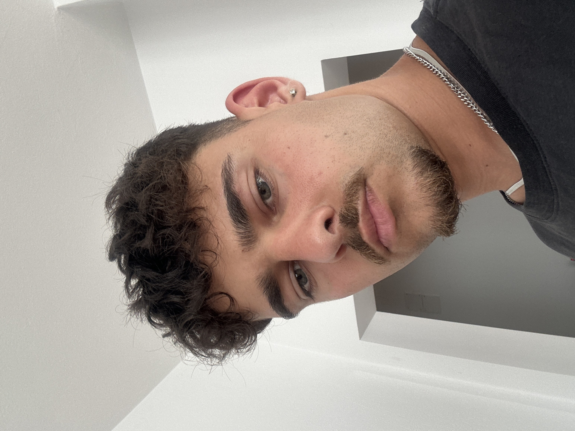 A portrait of a light-skinned man with dark curly hair, a beard, and gray eyes. He is wearing a dark gray t-shirt and a silver chain necklace. The background is a plain white wall with a shadowed corner of the ceiling visible. The photo is taken at a low angle, looking up at the subject. There is no indication of Troyan, Bulgaria.